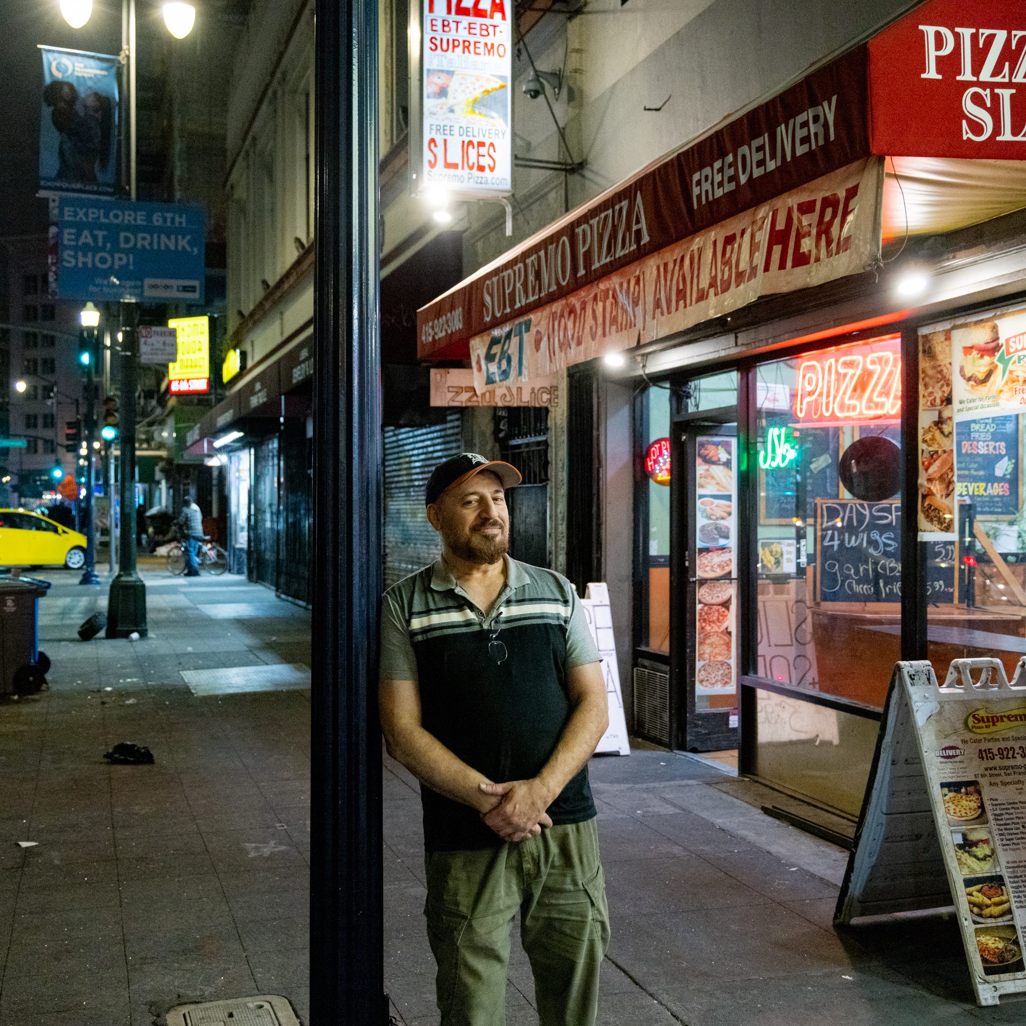 A man stands under a street lamp at night in front of &quot;Supremo Pizza.&quot; The street is lined with various storefronts, neon signs, and advertisements for pizza slices.