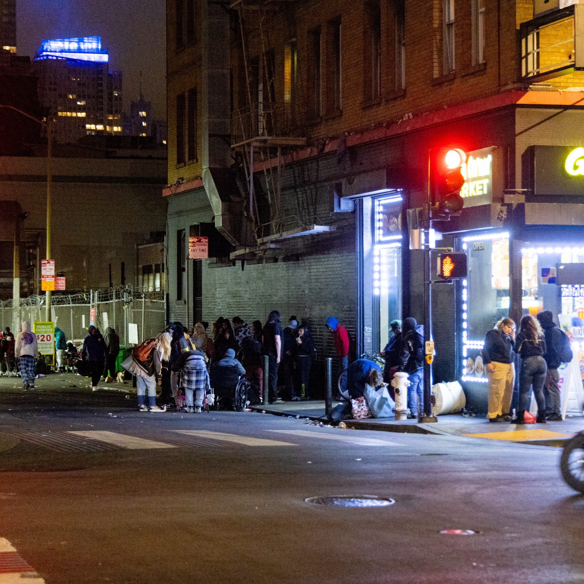 People gather outside a brightly lit market on a city street at night, some standing while others sit or lean against the building, with skyscrapers visible in the background.