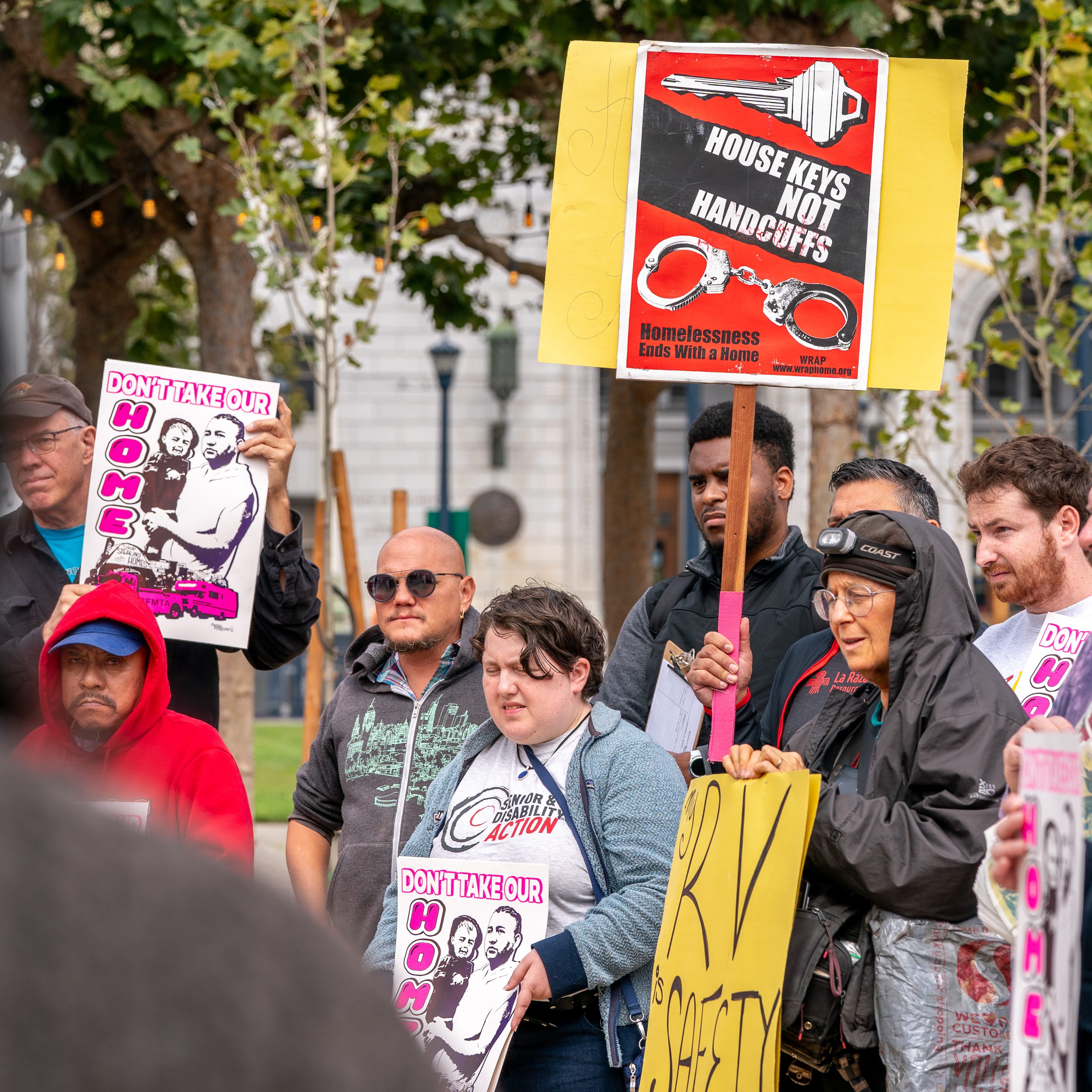 A group of people is holding various signs at a protest. One sign says, &quot;HOUSE KEYS NOT HANDCUFFS,&quot; another displays &quot;DON'T TAKE OUR HOME&quot; with an image of a family.