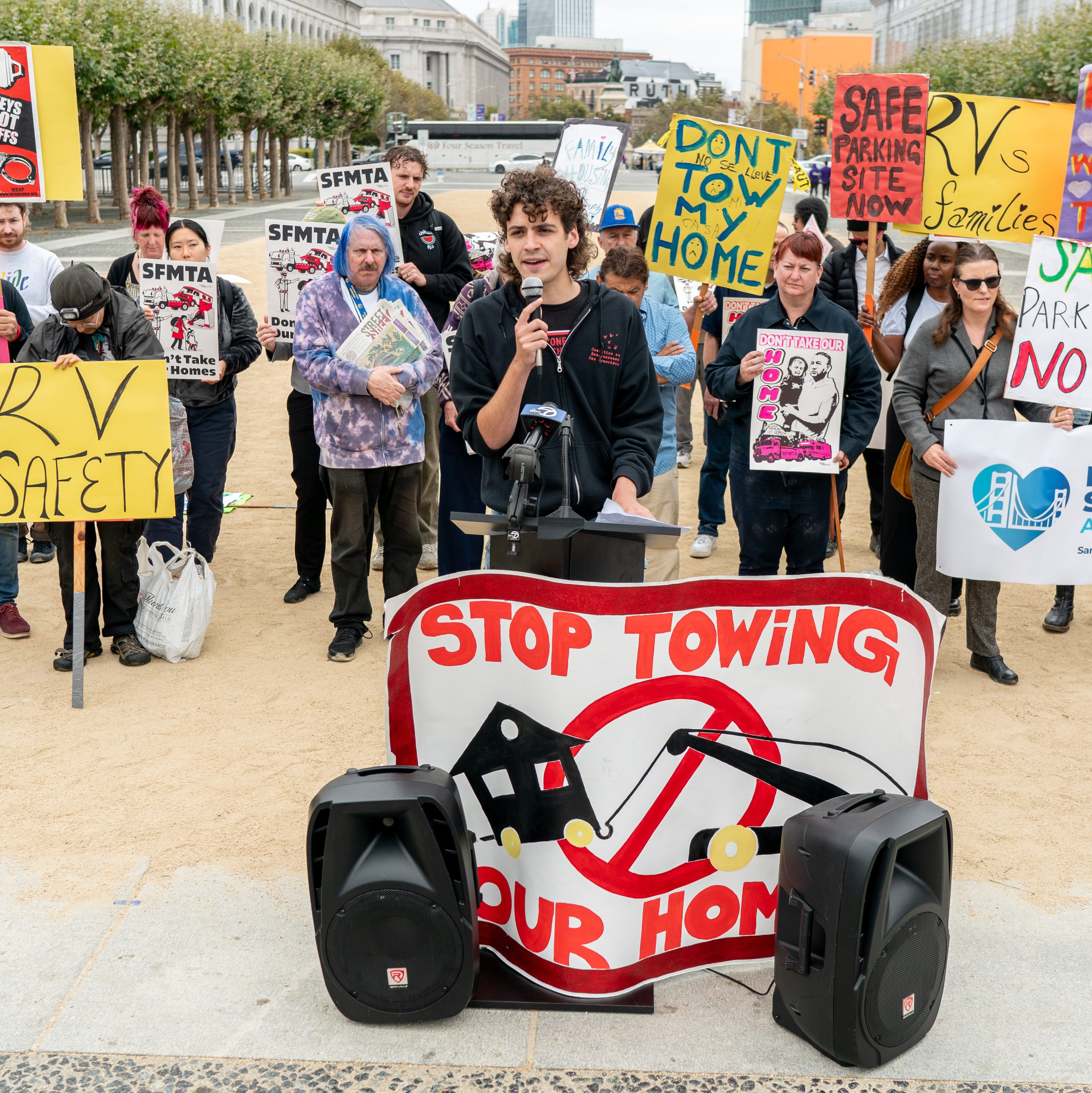 A man speaks at a podium surrounded by people holding signs protesting RV towing. Prominent signs include messages like &quot;RV Safety,&quot; &quot;Don't Tow My Home,&quot; and &quot;Stop Towing Our Homes.&quot;