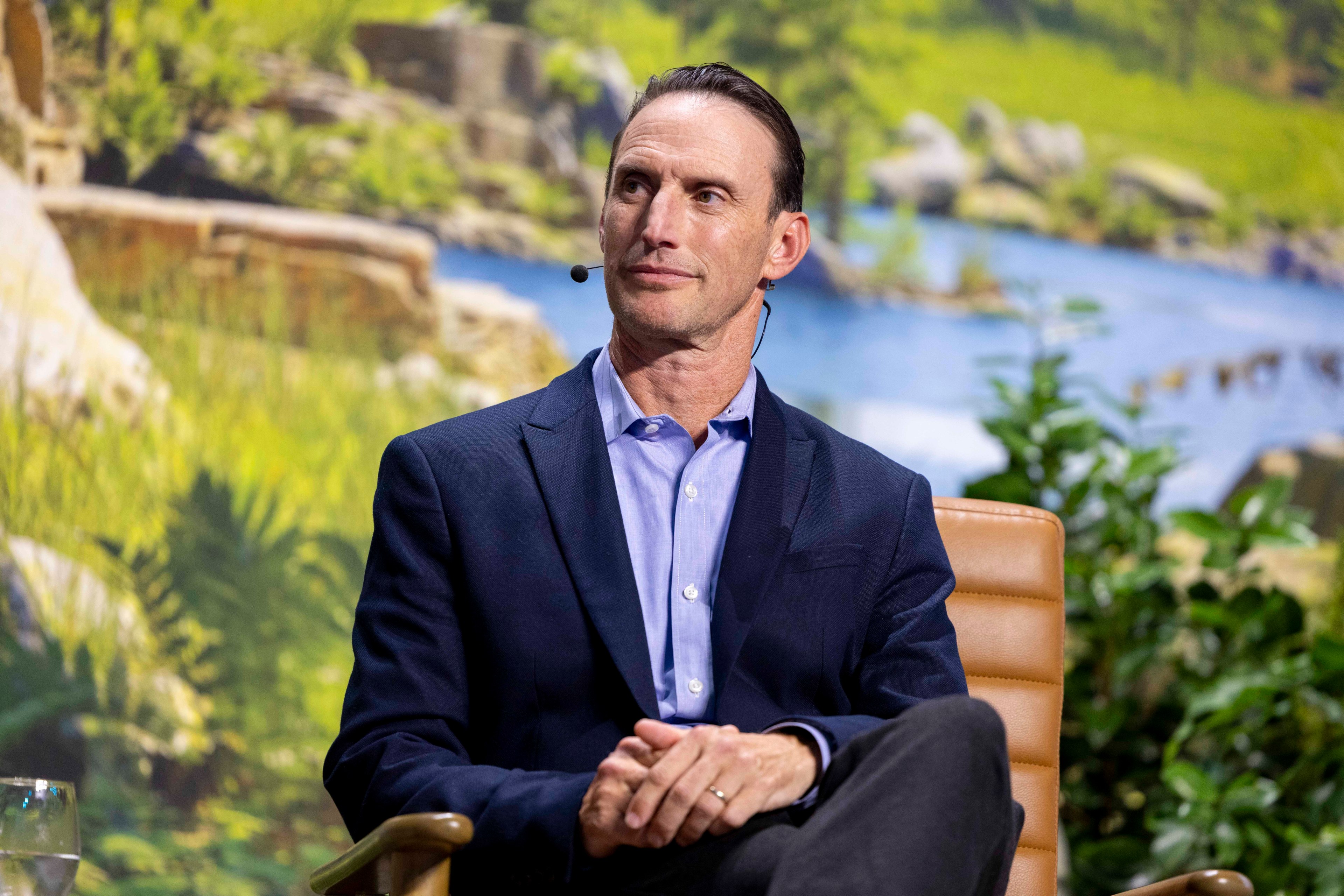 A man in a blue suit sits on a tan chair with a headset microphone, framed by a bright, nature-themed background with greenery and a flowing river.