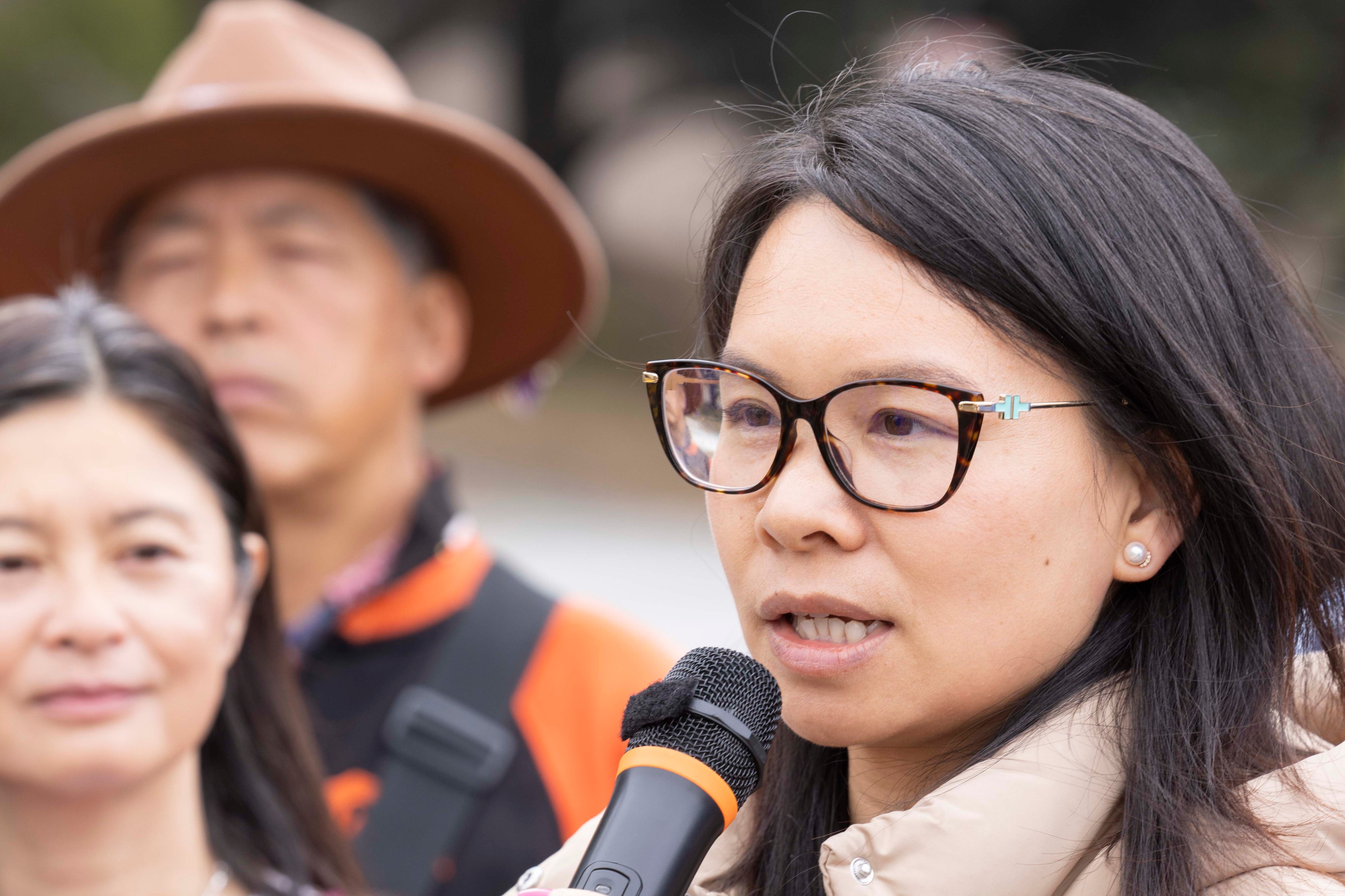 A woman in glasses speaks into a microphone, wearing a beige jacket. Behind her, another woman and a man in a hat stand, holding a blue sign with partial text.