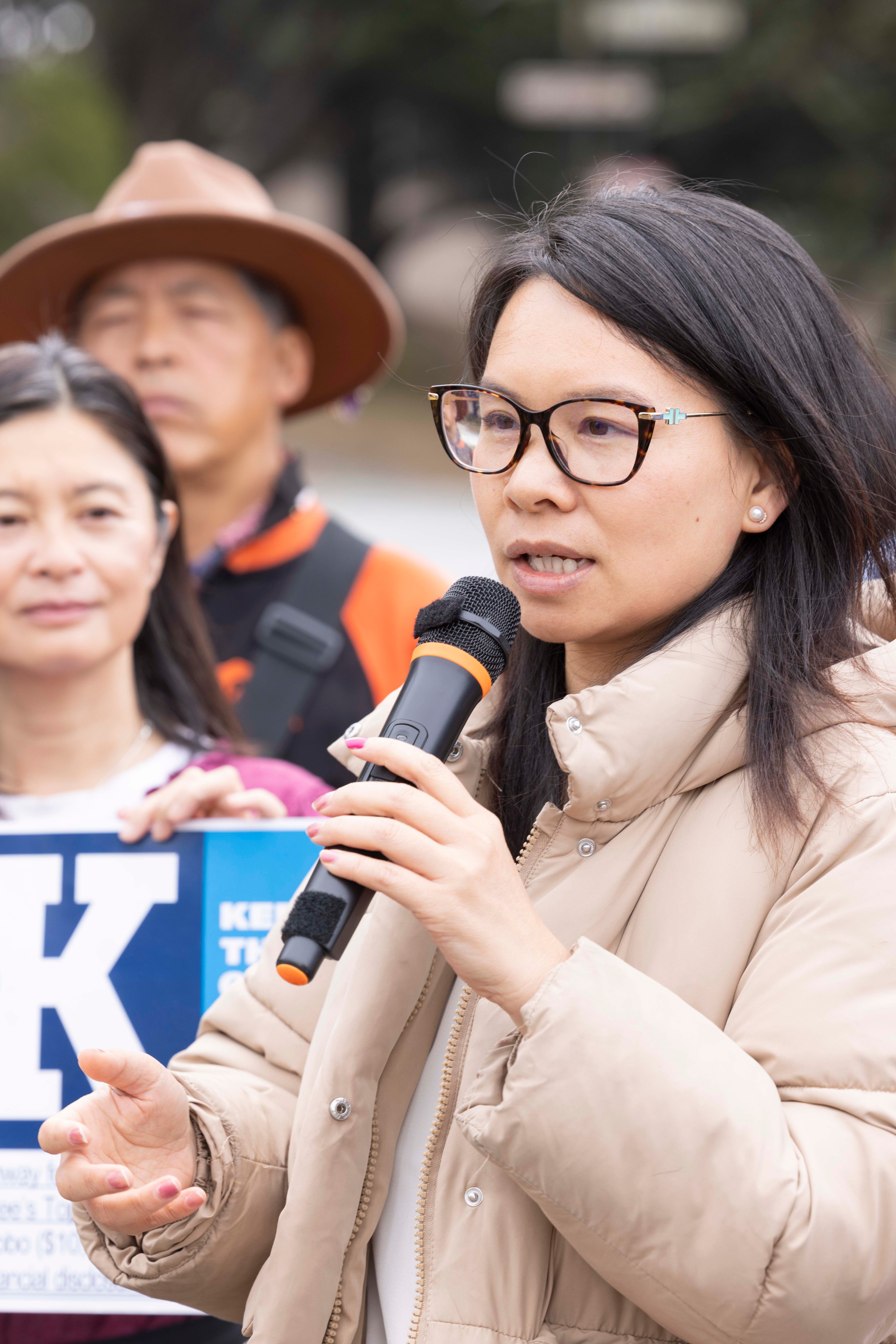 A woman in glasses speaks into a microphone, wearing a beige jacket. Behind her, another woman and a man in a hat stand, holding a blue sign with partial text.