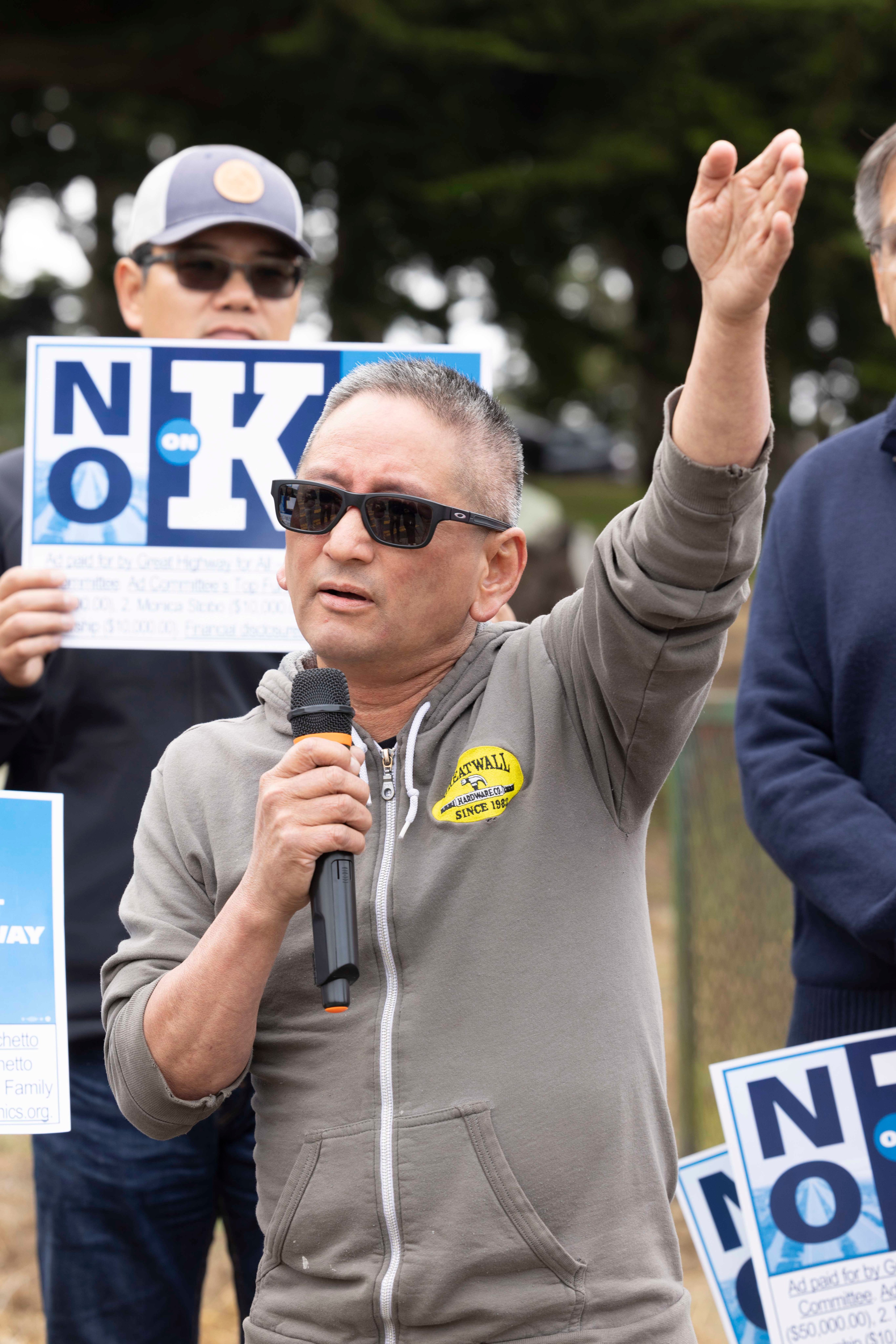 A man in a gray hoodie, speaking into a microphone with one hand raised, stands outdoors in front of a group. He is wearing sunglasses, and others hold &quot;No on K&quot; signs.