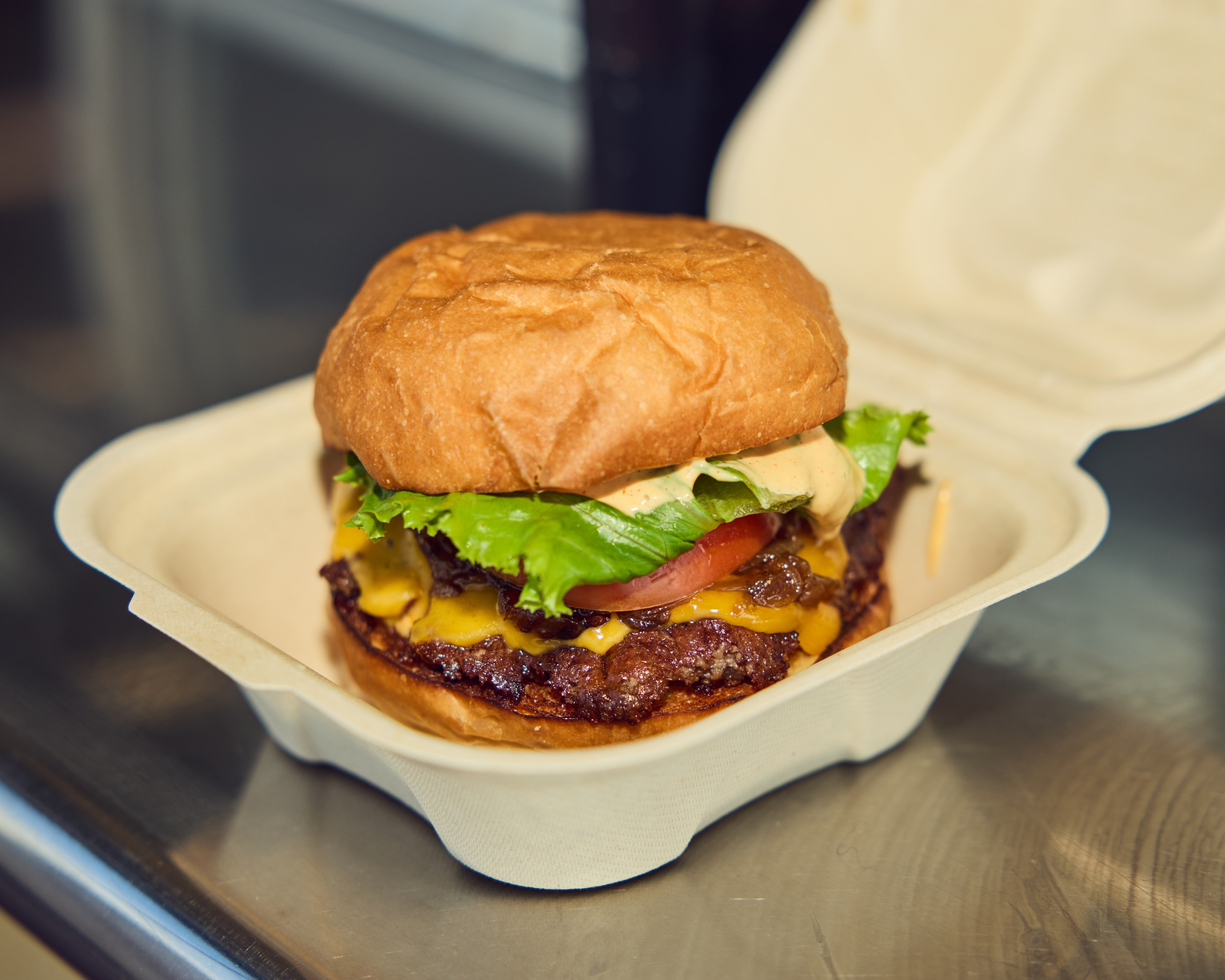 The image shows a cheeseburger with a beef patty, lettuce, tomato, melted cheese, and sauce inside a toasted bun, placed in an open takeout container.