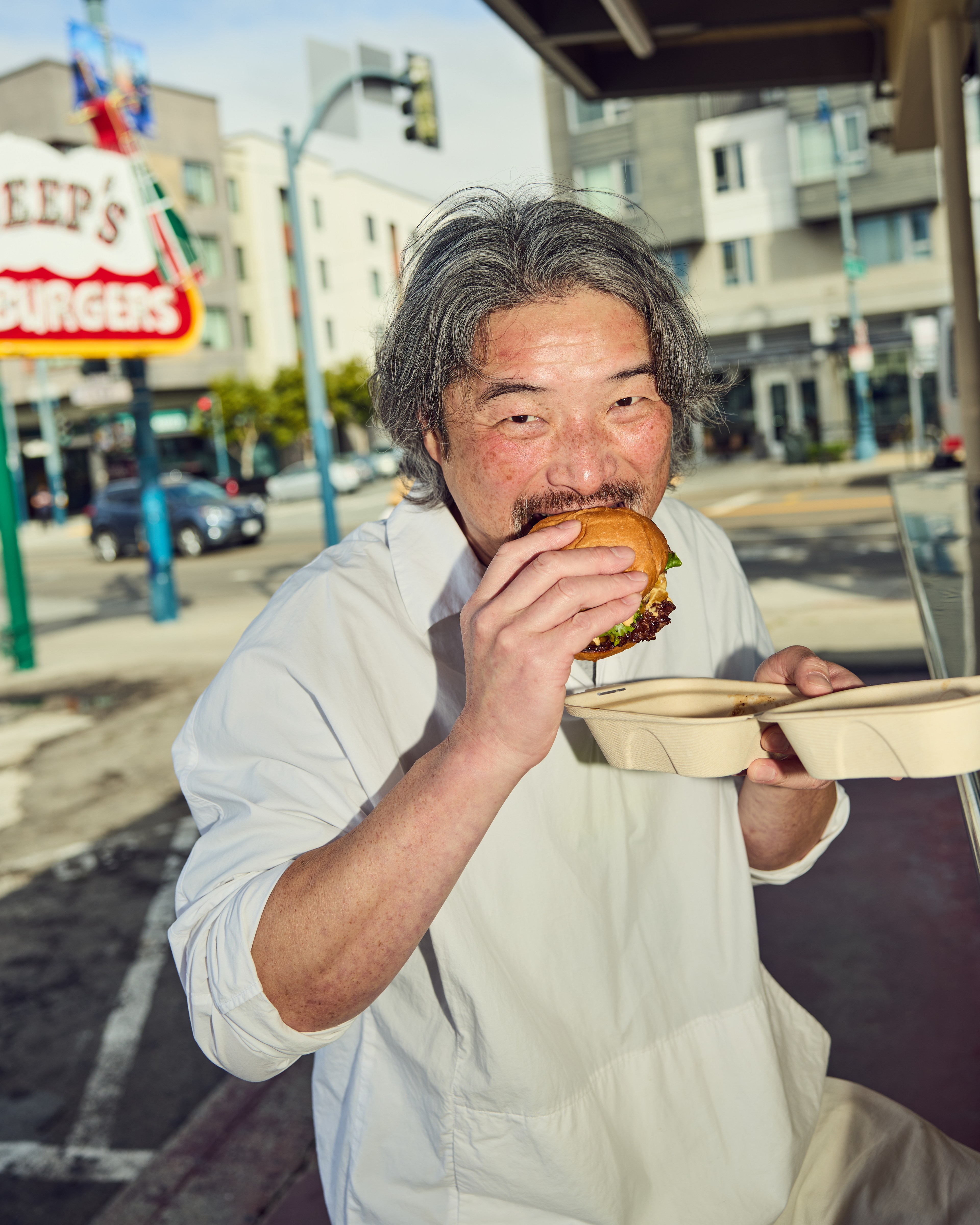 A man with grey hair is enjoying a large burger while standing outside, holding a takeout container. A street and buildings are visible in the background.