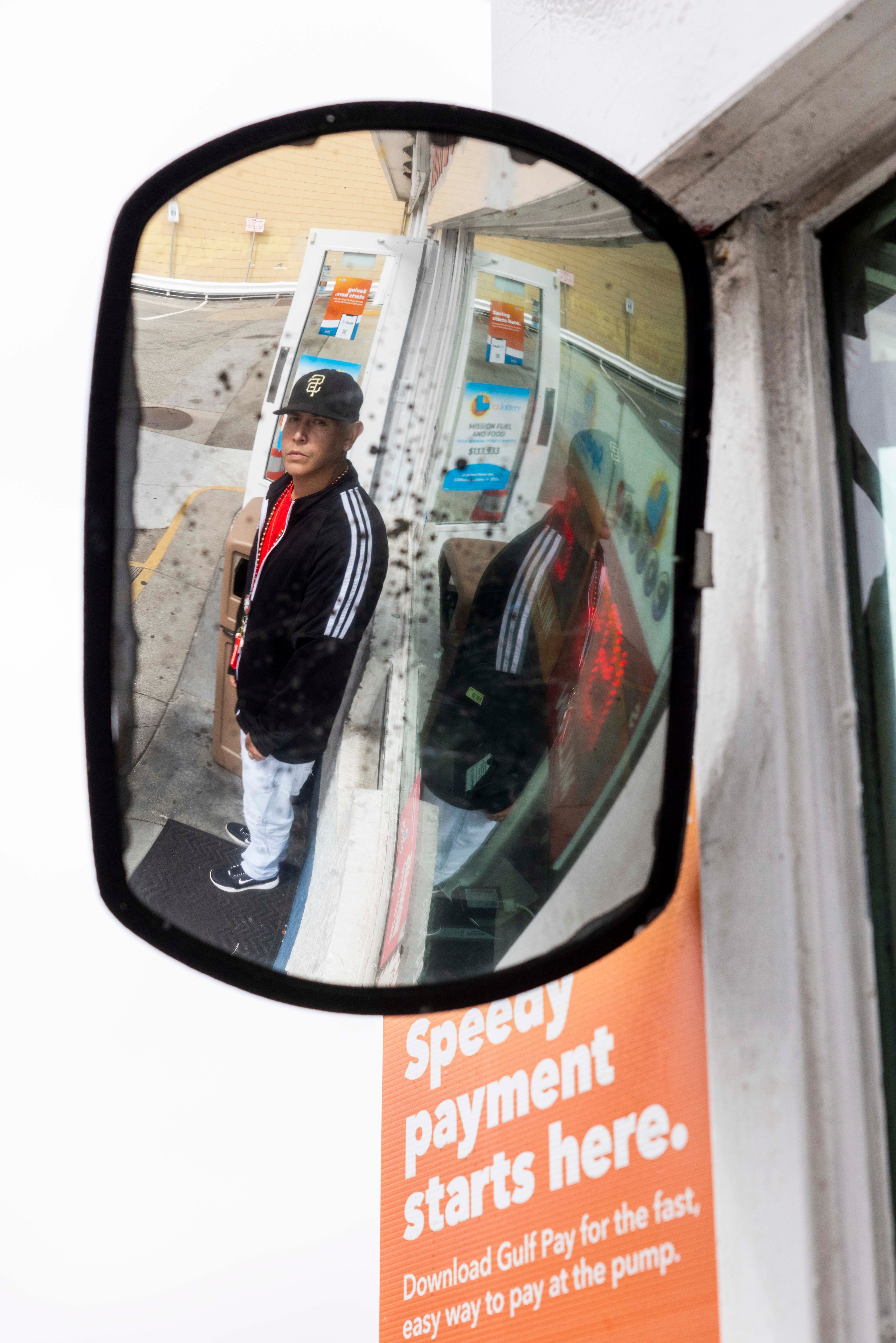 A man is seen reflected in a dirty convex mirror, standing near a gas station window with signage about speedy Gulf Pay payments. He wears a black cap and jacket.