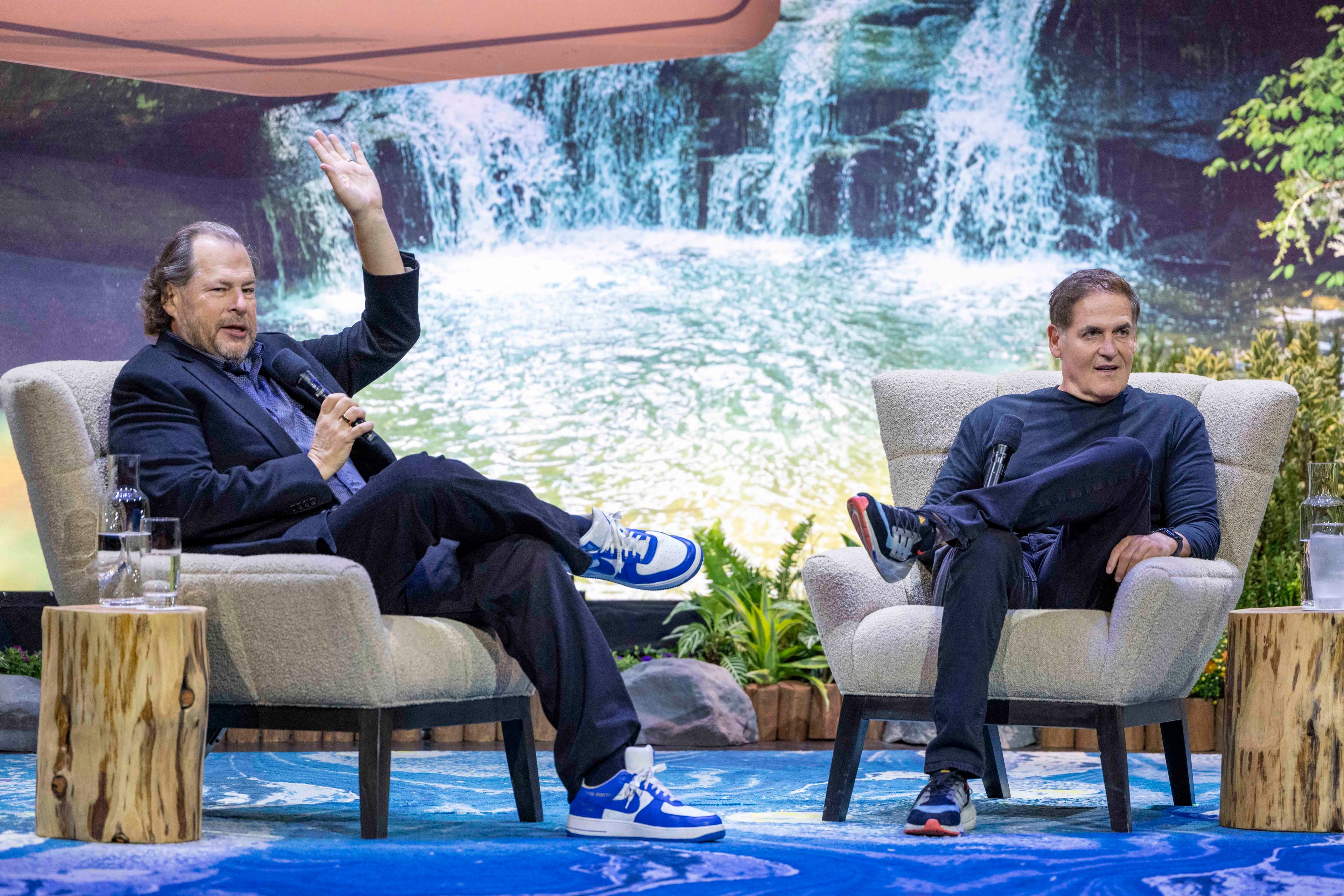 Two men sit in armchairs on a stage with a waterfall backdrop. One, in a suit and blue sneakers, raises his hand while another, in casual attire, looks at him.