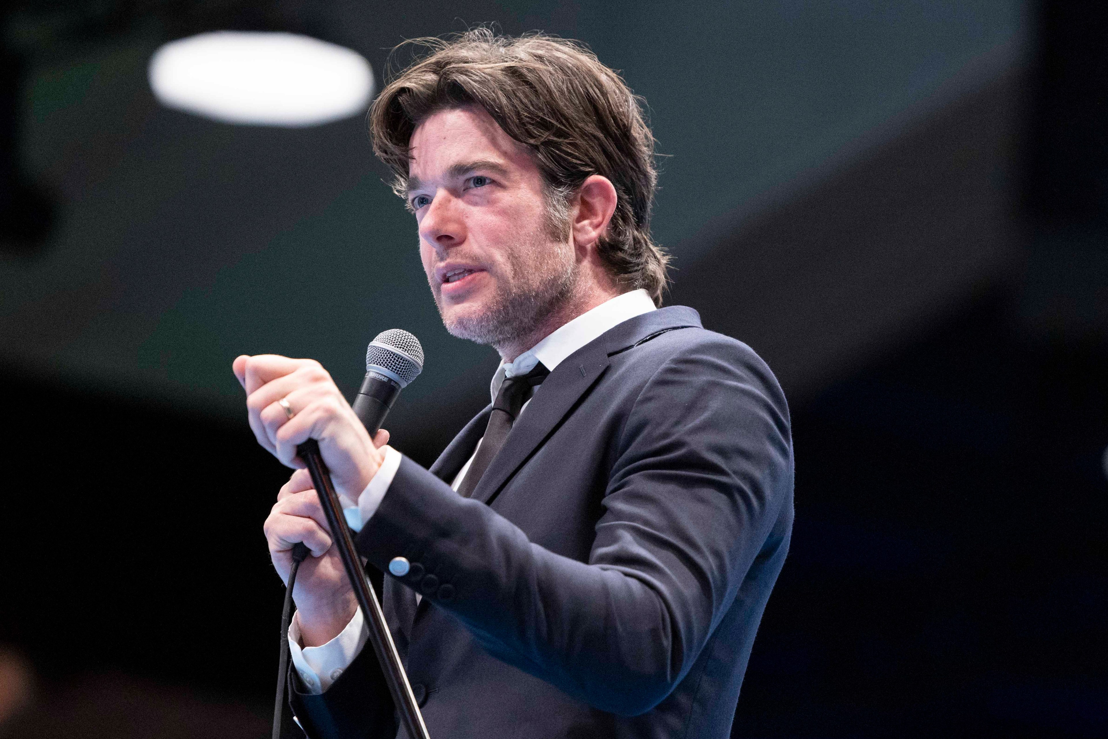 A man in a navy suit and black tie is holding a microphone and speaking on stage. The background is dimly lit, focusing attention on him.