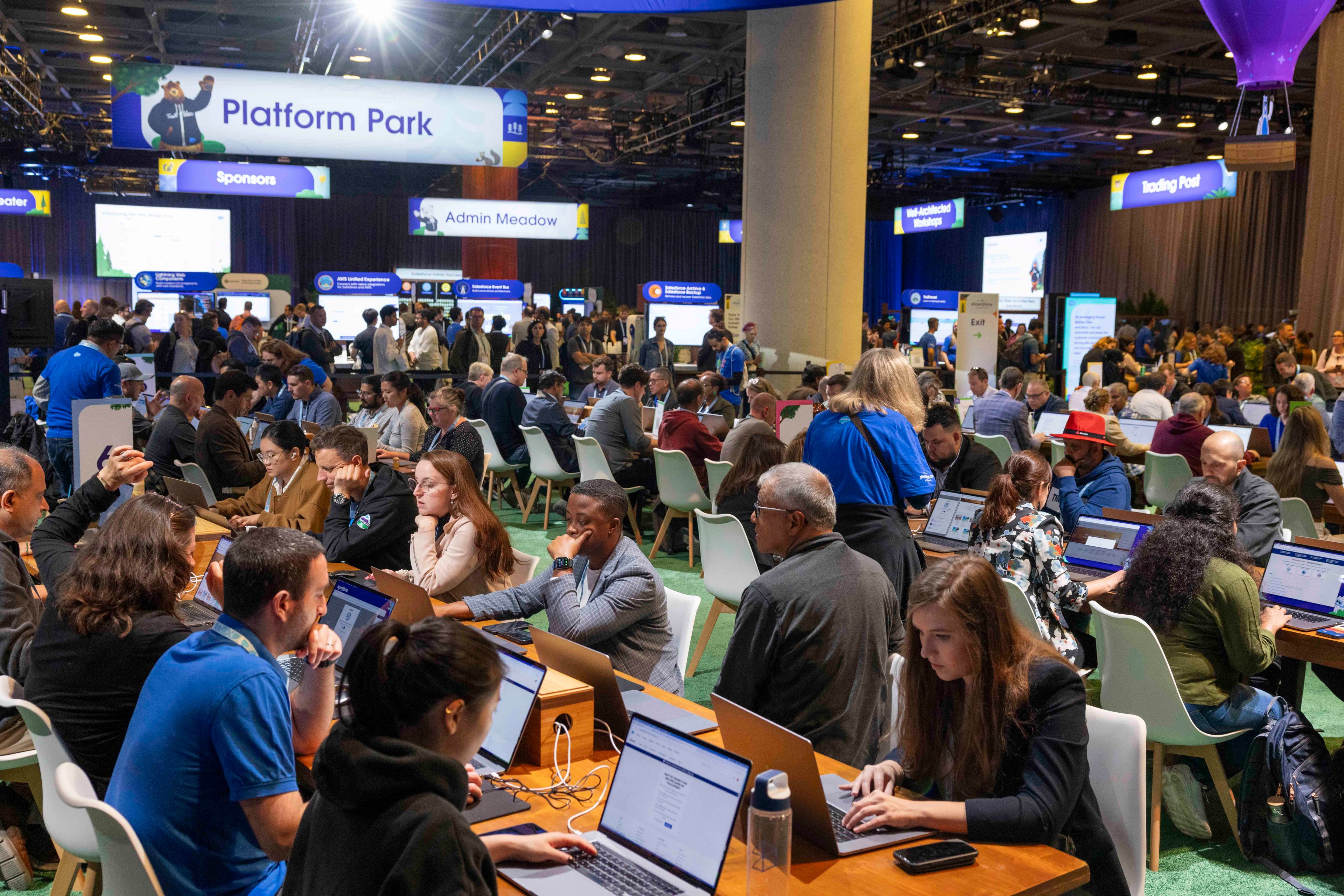 People fill a tech conference hall, working on laptops at long tables under signs reading &quot;Platform Park&quot; and &quot;Admin Meadow,&quot; and other designated areas.