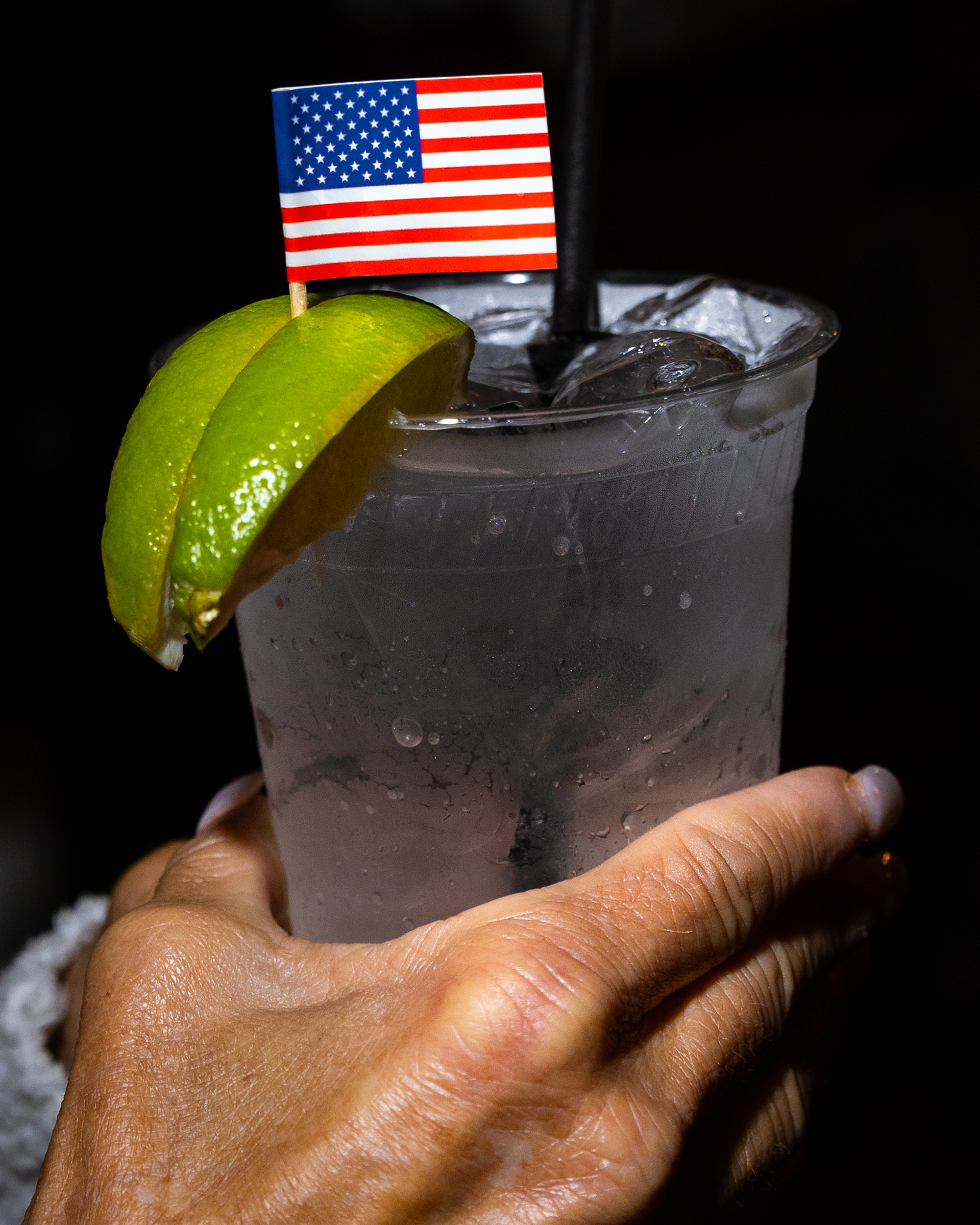 A hand holds a cup filled with a clear ice-filled drink garnished with lime wedges and a small American flag on a toothpick.