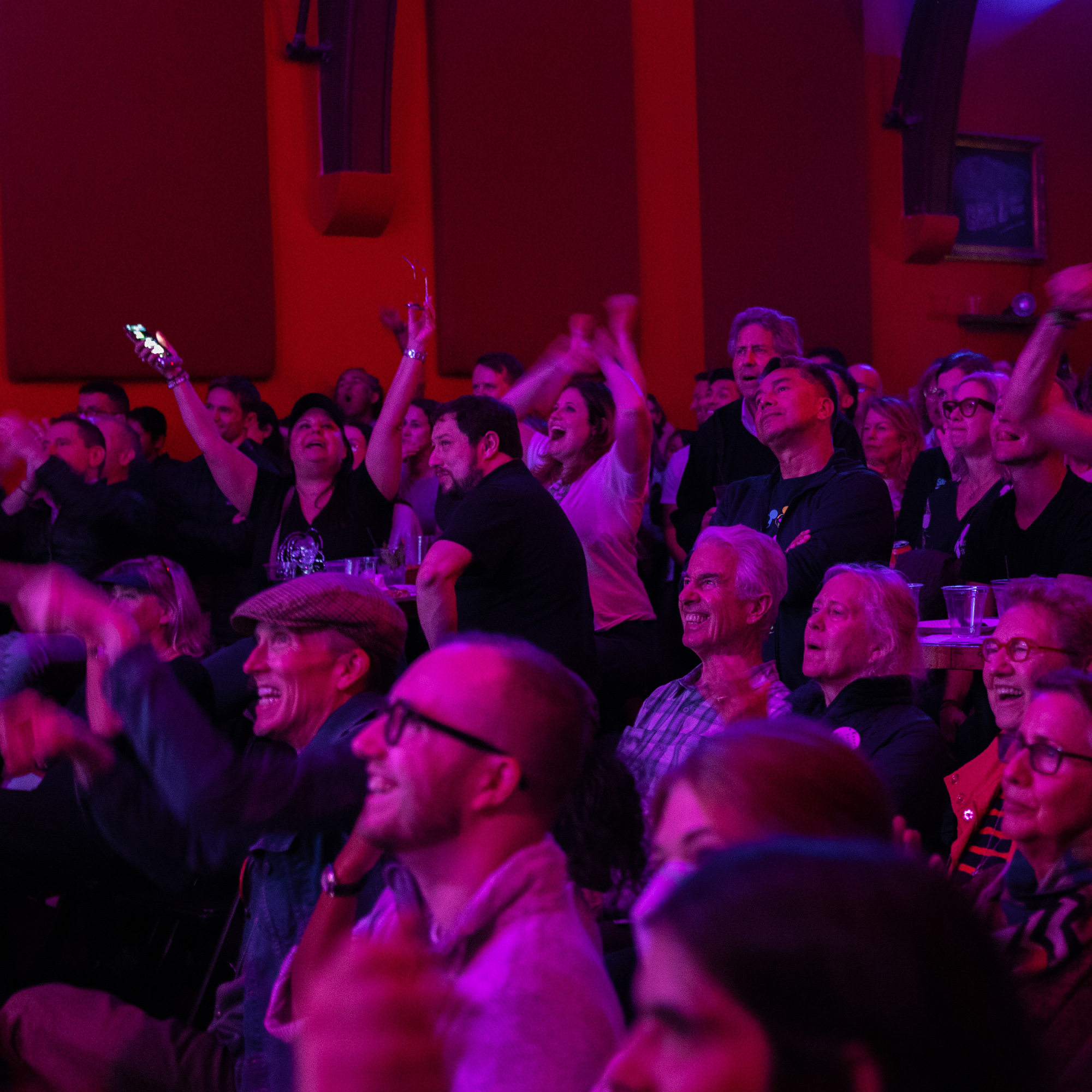 A crowd inside a room cheers with raised arms. The atmosphere is lively, with people displaying various expressions of excitement and joy against a reddish-purple backdrop.