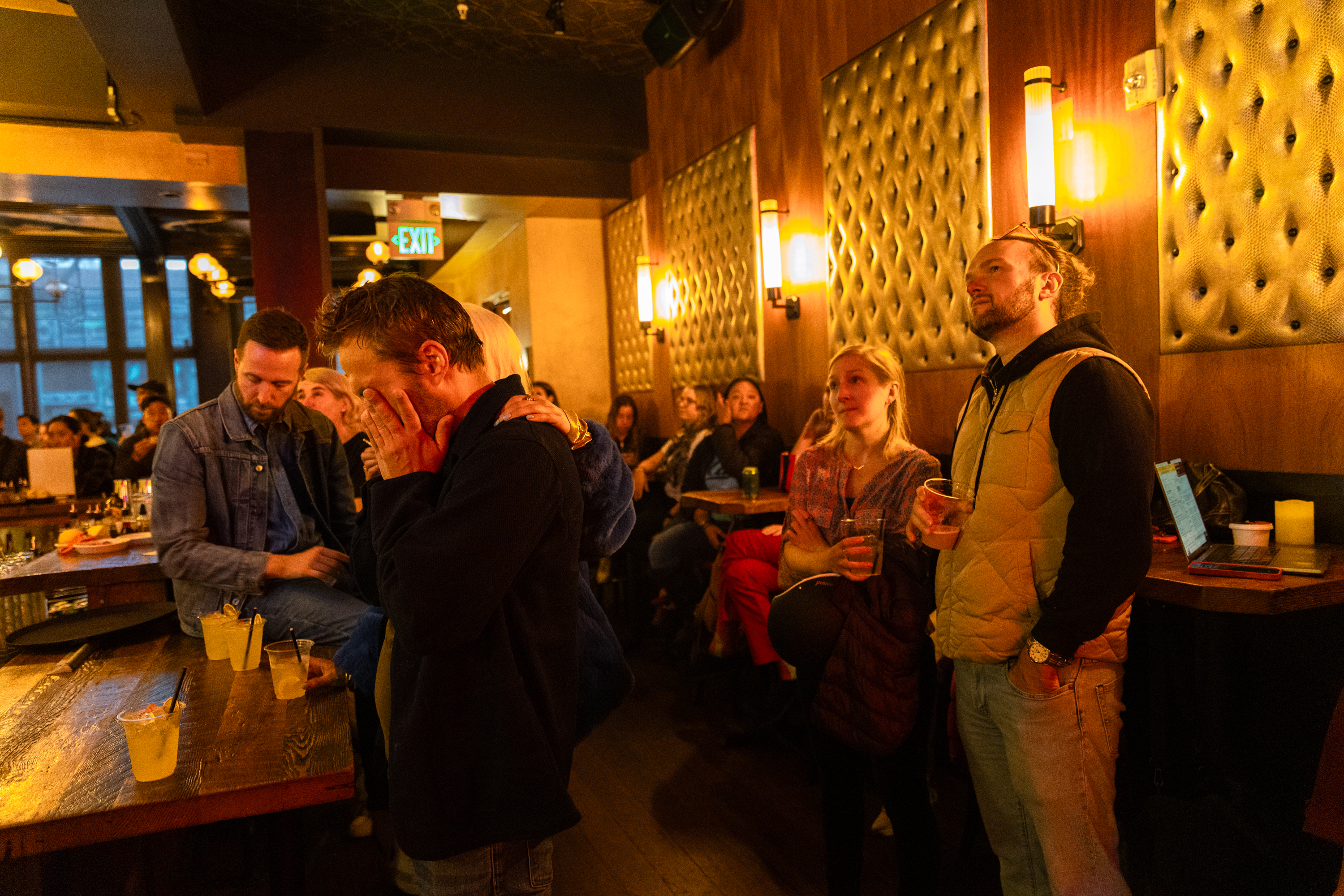 A group of people is in a dimly lit bar, their expressions ranging from concerned to distressed. Drinks are on the table, and the atmosphere appears tense.