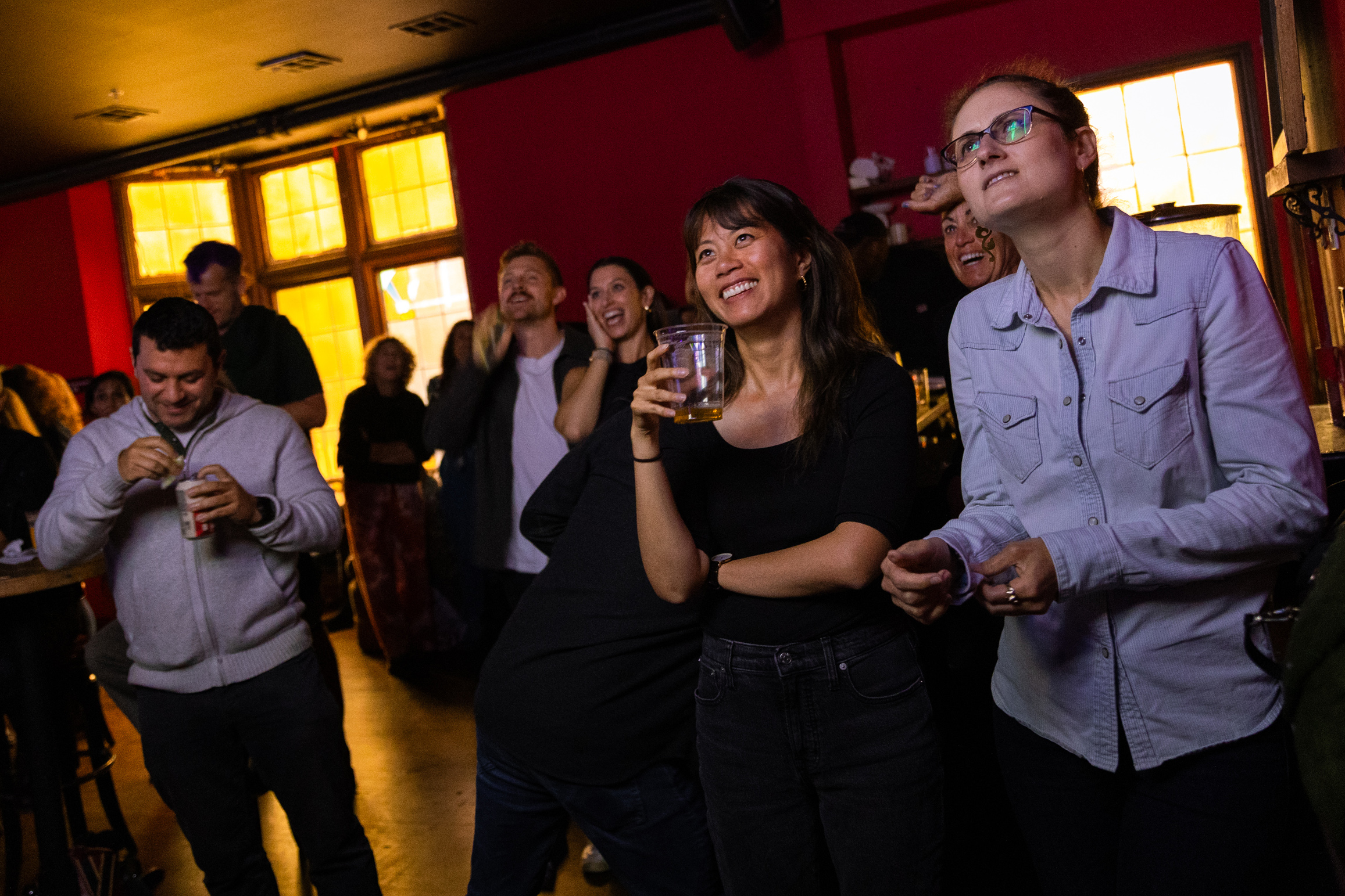 The image shows a group of people in a dimly lit room, watching something with expressions of joy and amusement. Some hold drinks, and large windows are in the background.