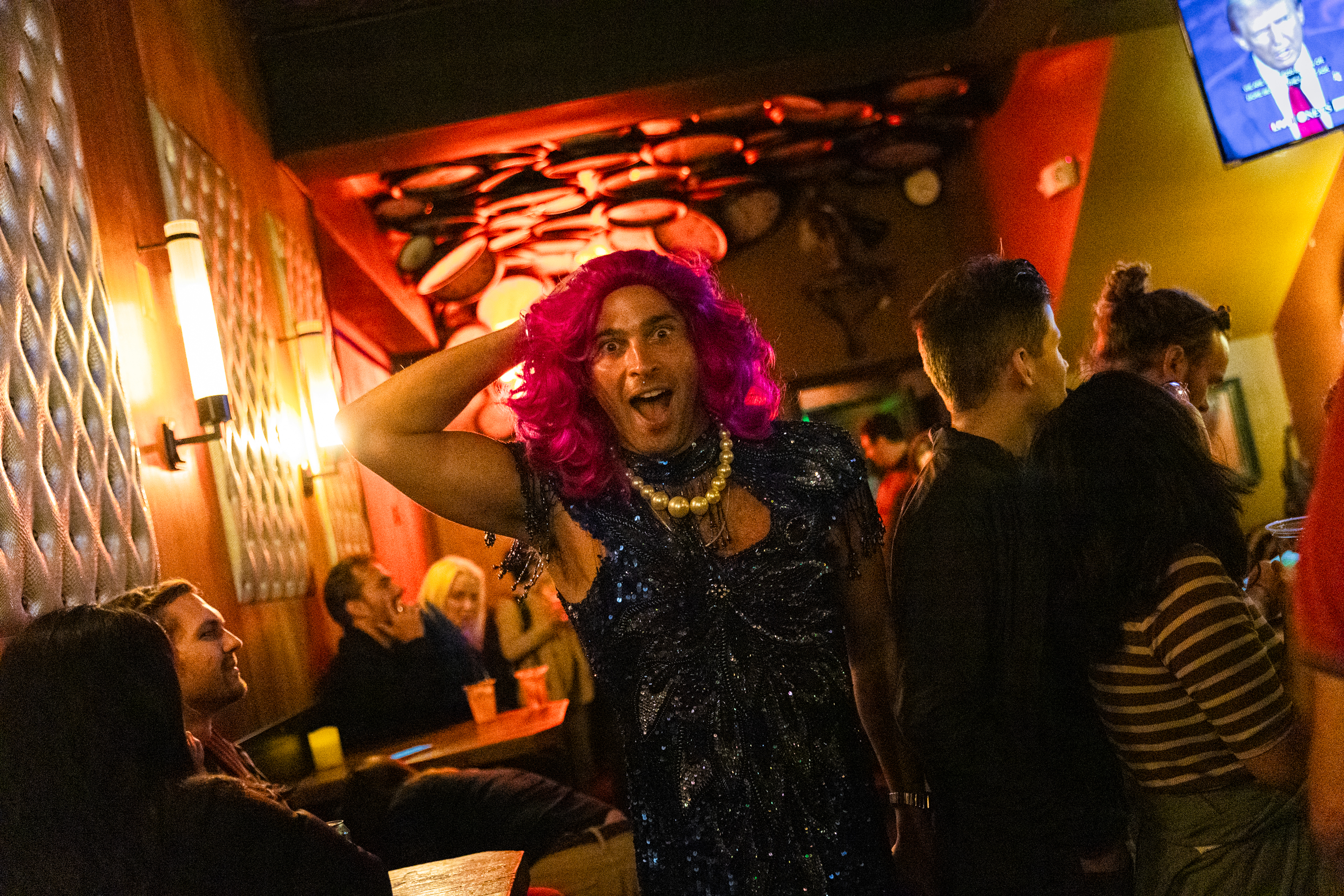 A lively person in a blue sequined dress and bright pink wig poses playfully in a dimly lit, vibrant bar, surrounded by chatting patrons and a TV in the background.