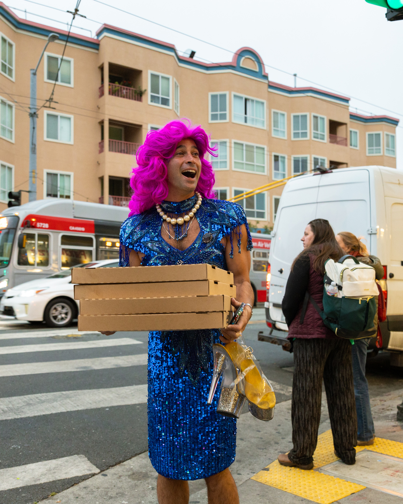 A person in a sparkling blue dress and vibrant pink wig stands at a crosswalk, holding pizza boxes and high-heeled shoes, with an urban street and buildings in the background.