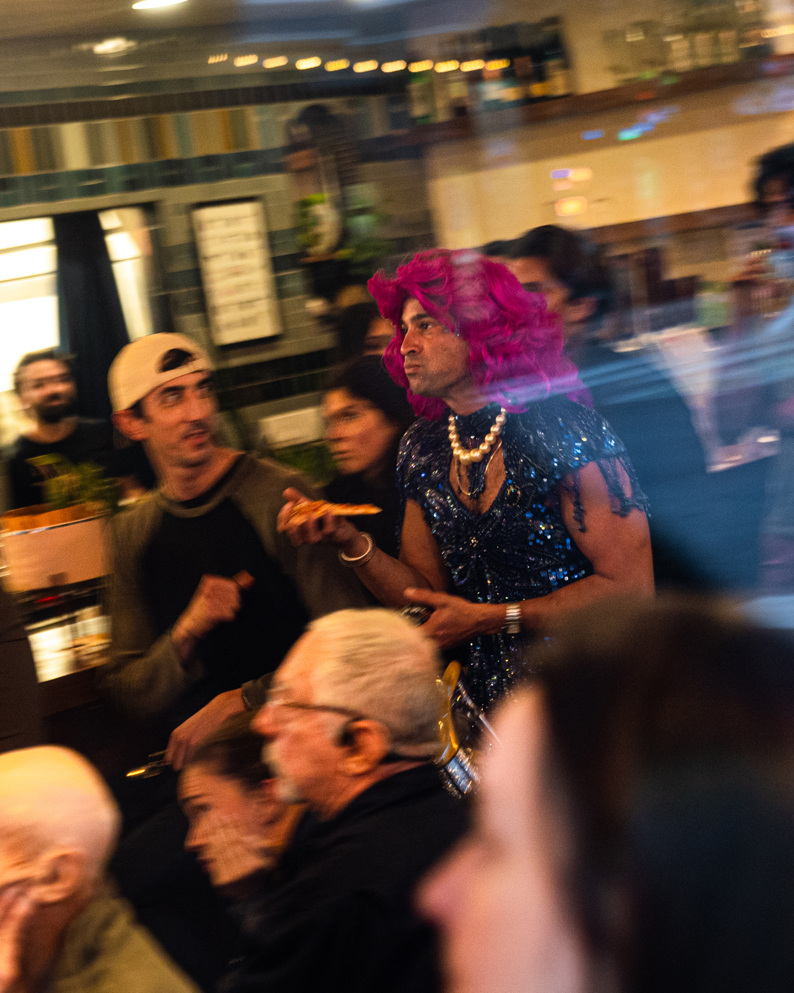 A person in a bright pink wig and sparkly dress stands among a crowd, holding a slice of pizza, with blurred surroundings suggesting a lively atmosphere.