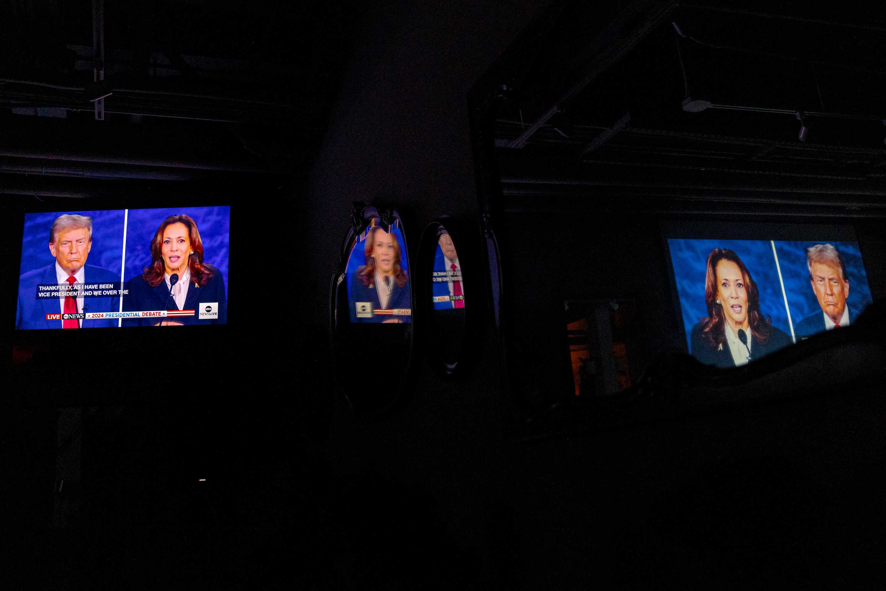 The image shows a televised debate between two political figures displayed on multiple screens and reflections in a dimly lit room.