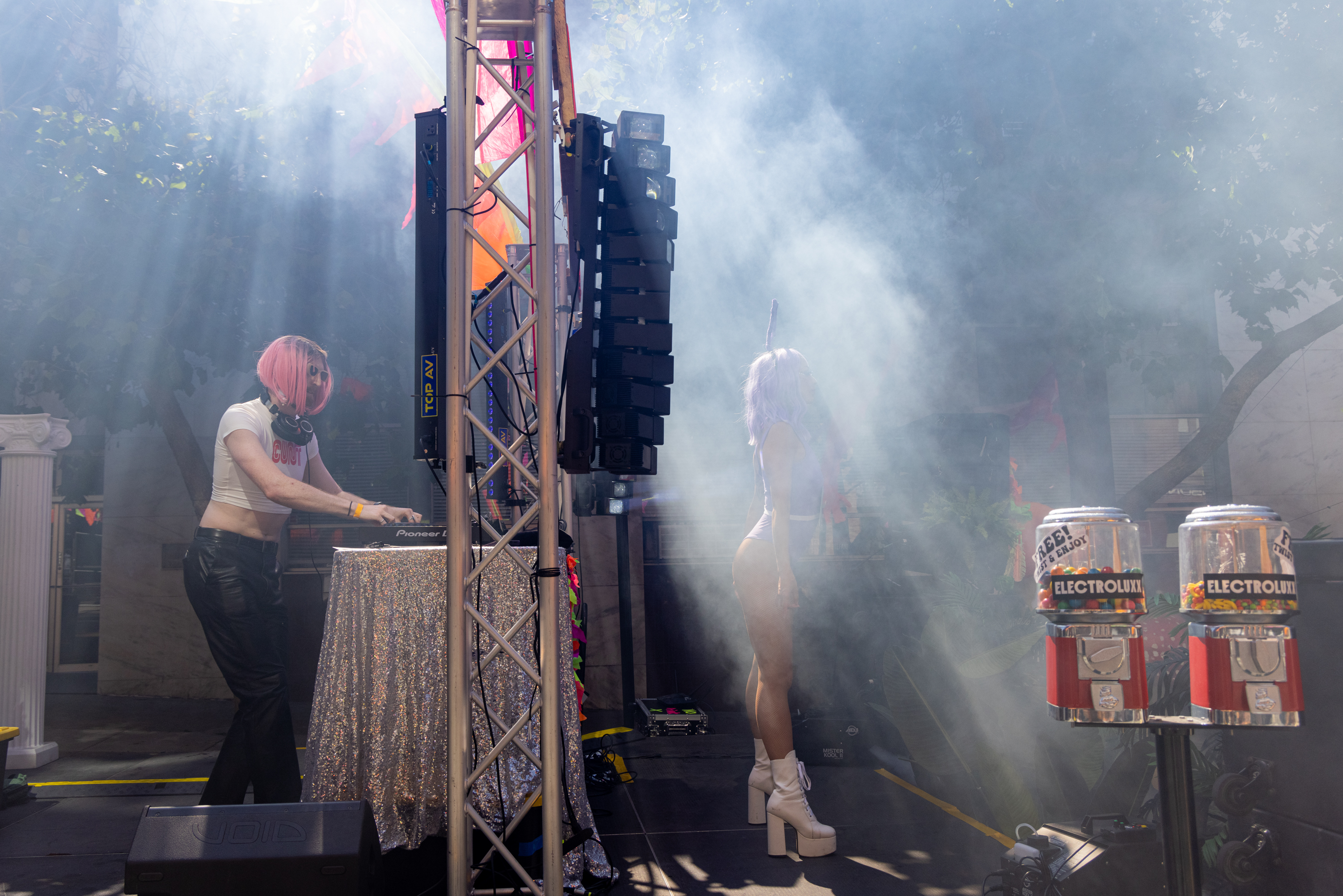 A DJ with pink hair and headphones performs on stage while a person with lavender hair and platform boots dances nearby, surrounded by colorful lights and machines.