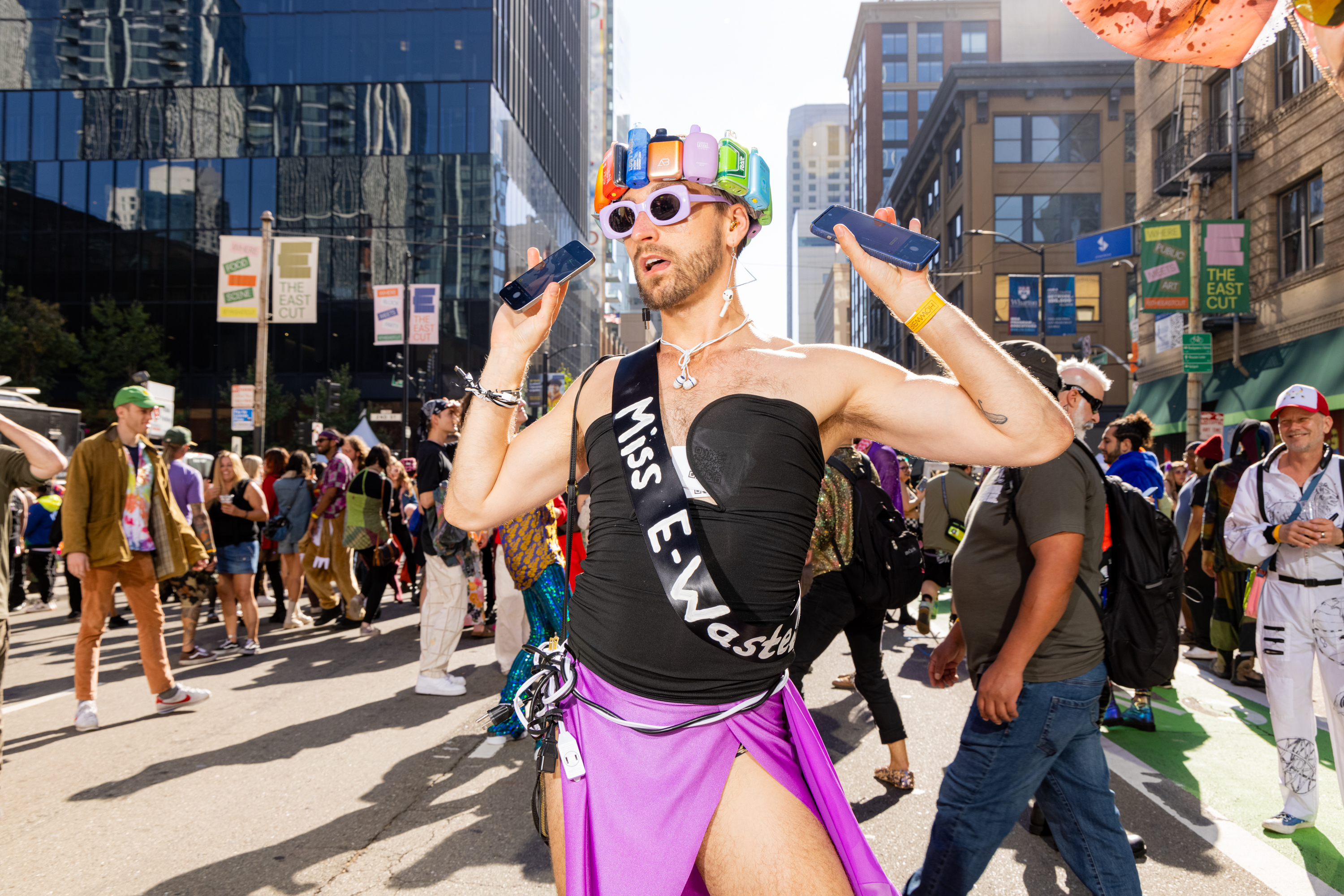 A person cheerfully holds two cell phones while wearing an electronic gadget hat, sunglasses, a &quot;Miss E-Waste&quot; sash, and a purple skirt amidst a busy street crowd.