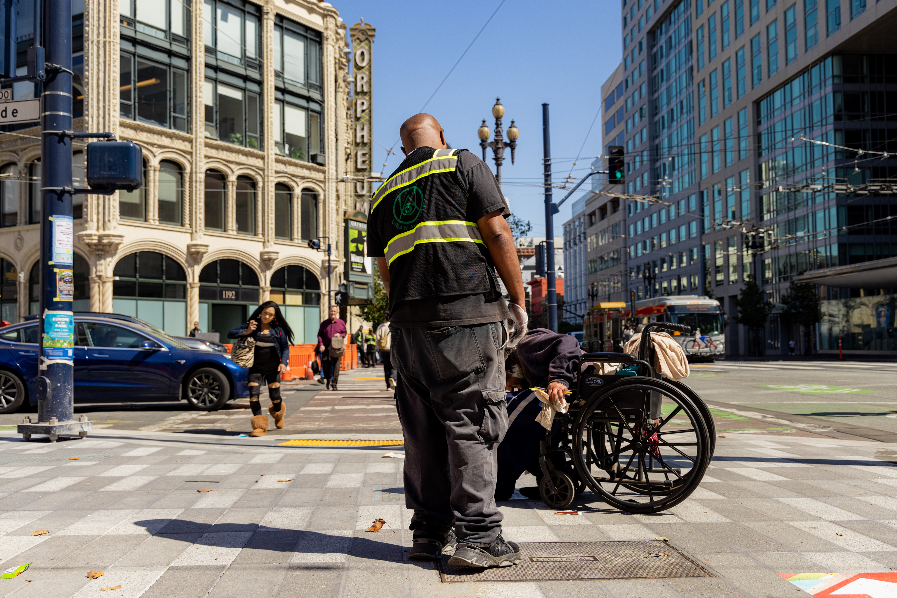 A man in a high-visibility vest bends down to assist someone in a wheelchair on a busy city sidewalk, surrounded by tall buildings, cars, and pedestrians.