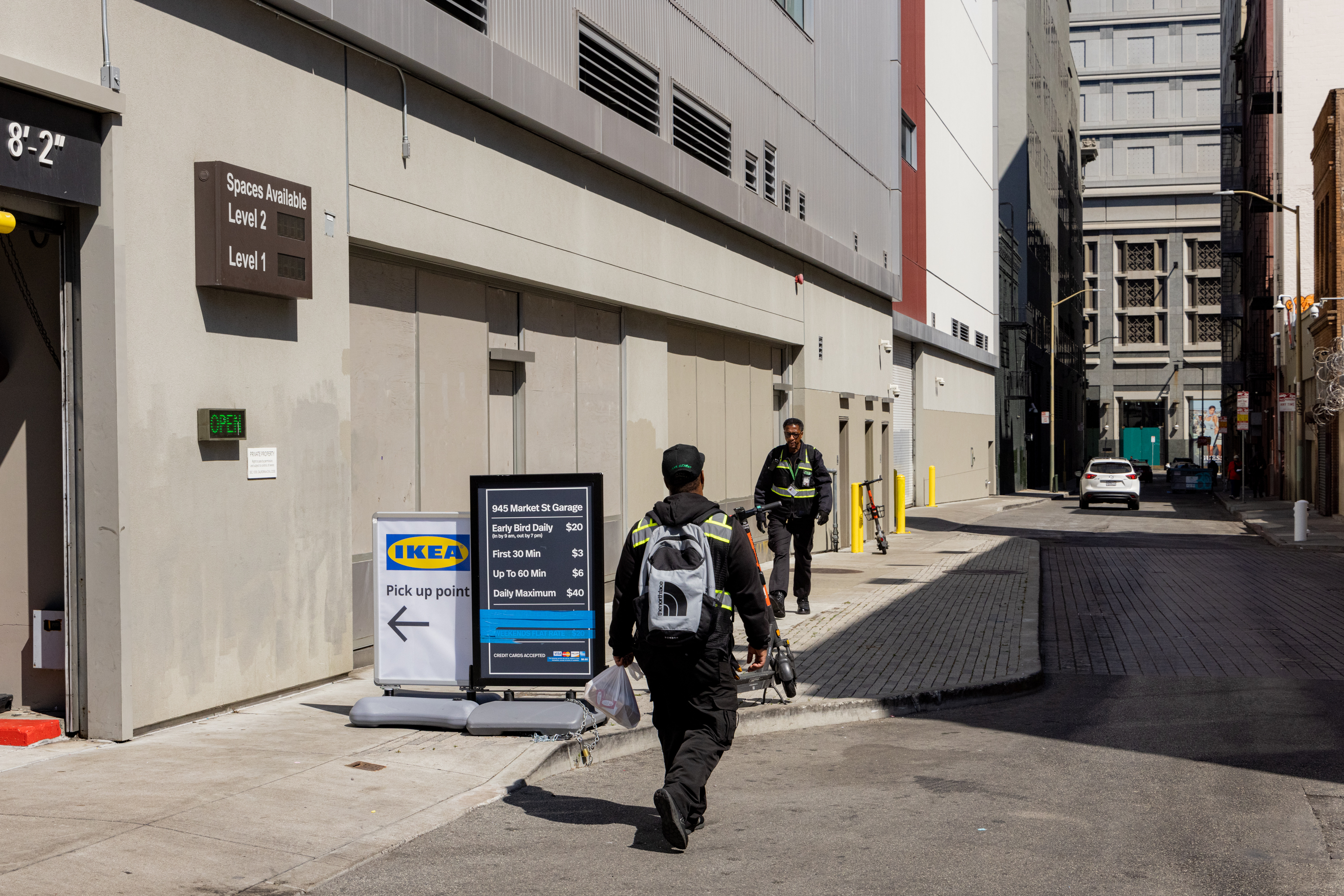 Two men, wearing black uniforms with reflective stripes, walk near a parking garage with signs for IKEA's pickup point and parking rates. A white car is in the distance.