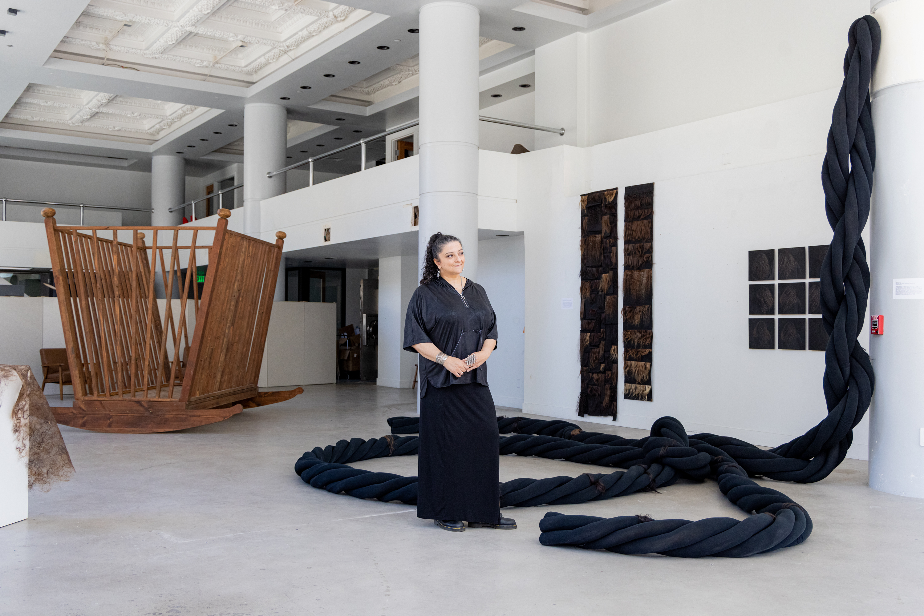 A woman stands in a minimalist, spacious art gallery with high ceilings. Behind her are large art installations, including a wooden structure and black twisted fabric.