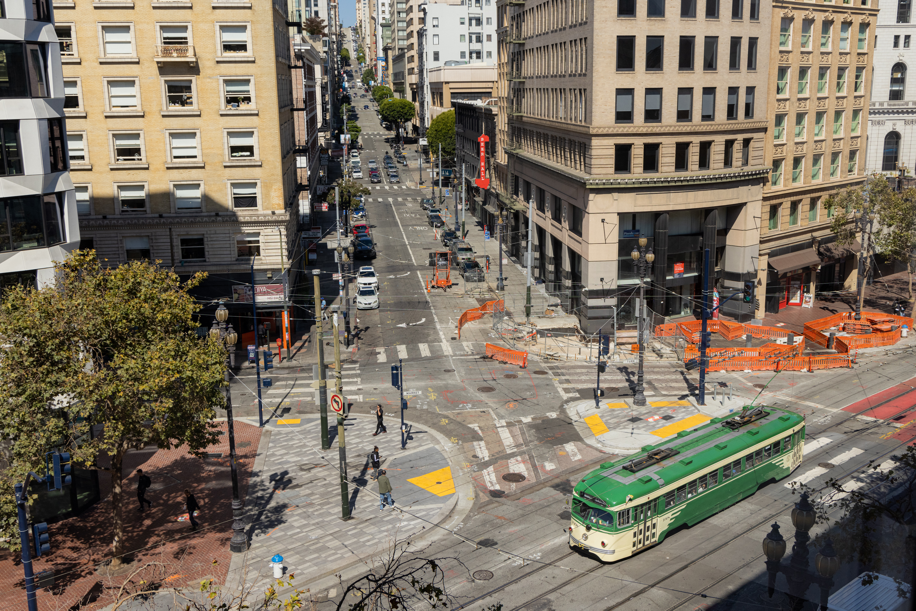 The image shows a city intersection with tall buildings, a green streetcar, pedestrians, cars, traffic signals, crosswalks, and orange construction barriers.