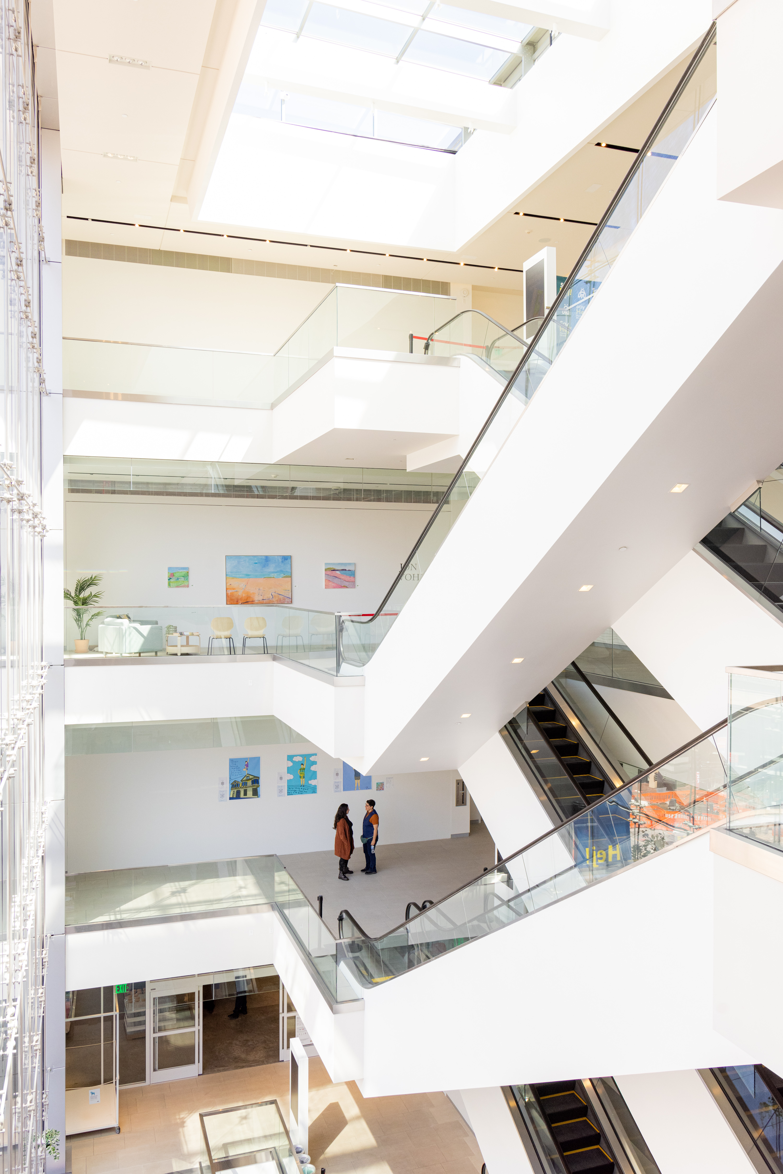 The image shows a bright, multi-level modern atrium with large windows, escalators, and people walking. Art is displayed on the walls, and natural light fills the space.