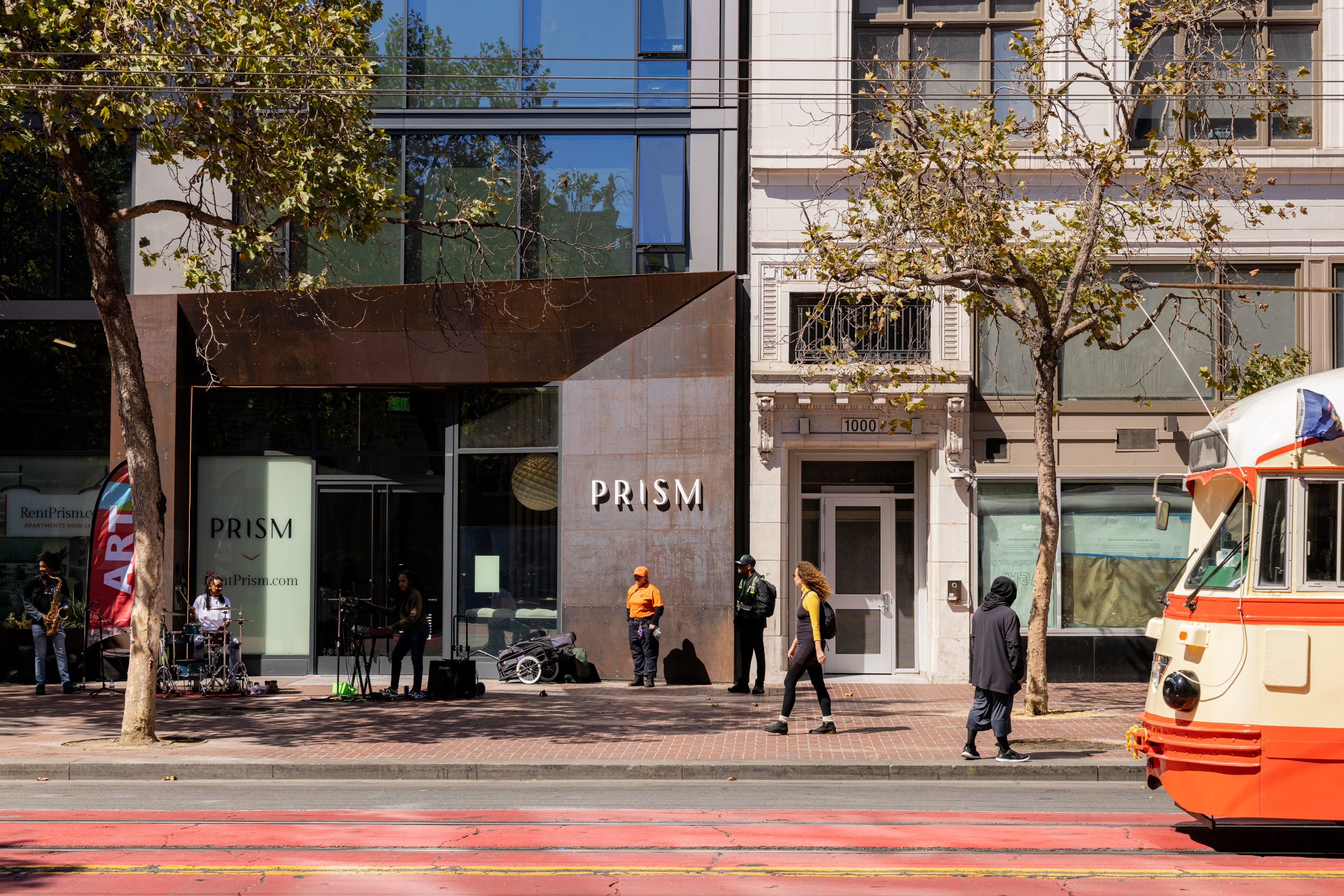 A street scene shows a building with the sign &quot;PRISM&quot; and a band playing outside. People walk by, and a vintage streetcar in cream and orange colors is passing.