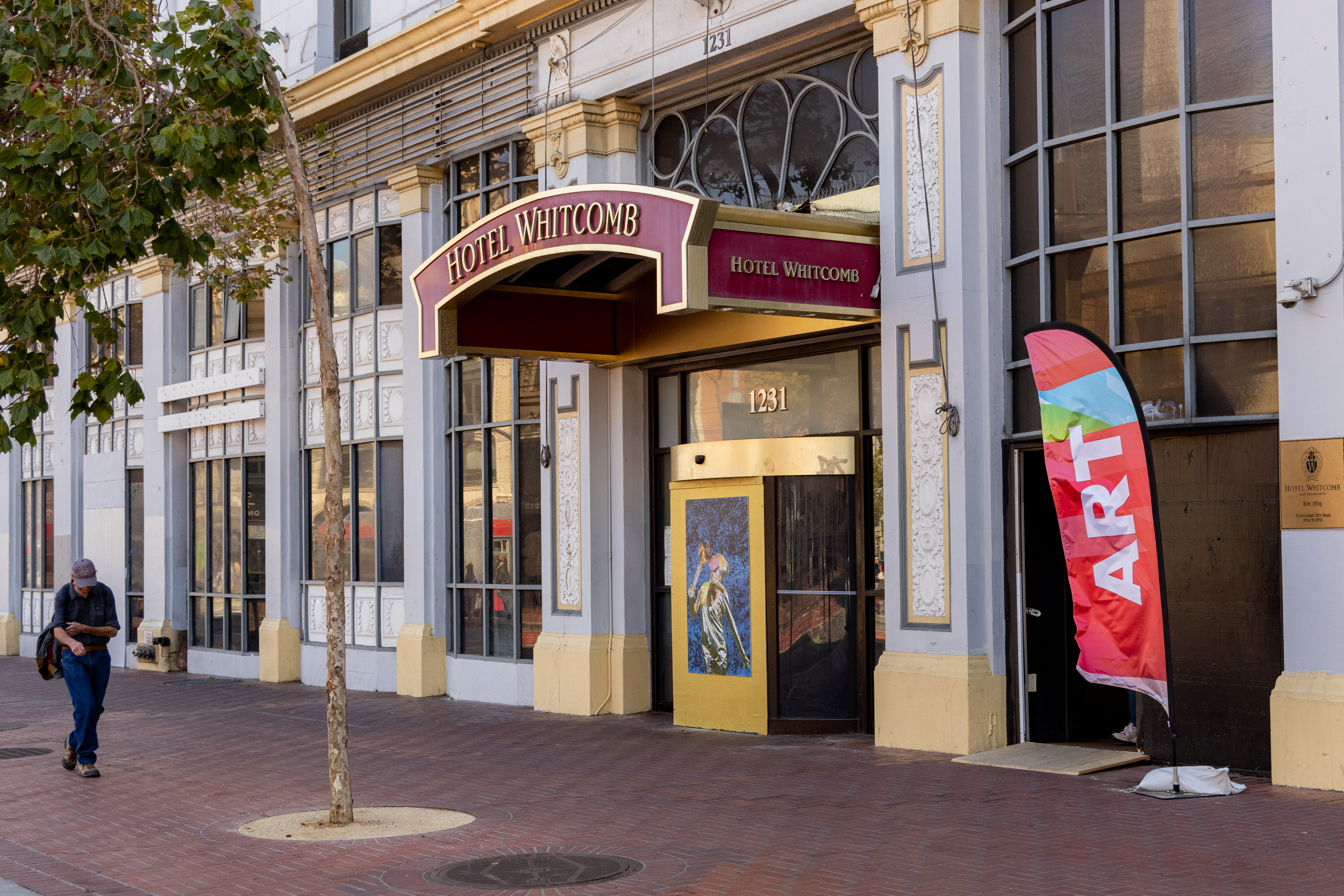 The image shows the entrance of Hotel Whitcomb with a red canopy, a colorful &quot;ART&quot; flag, and a person walking along the brick sidewalk.