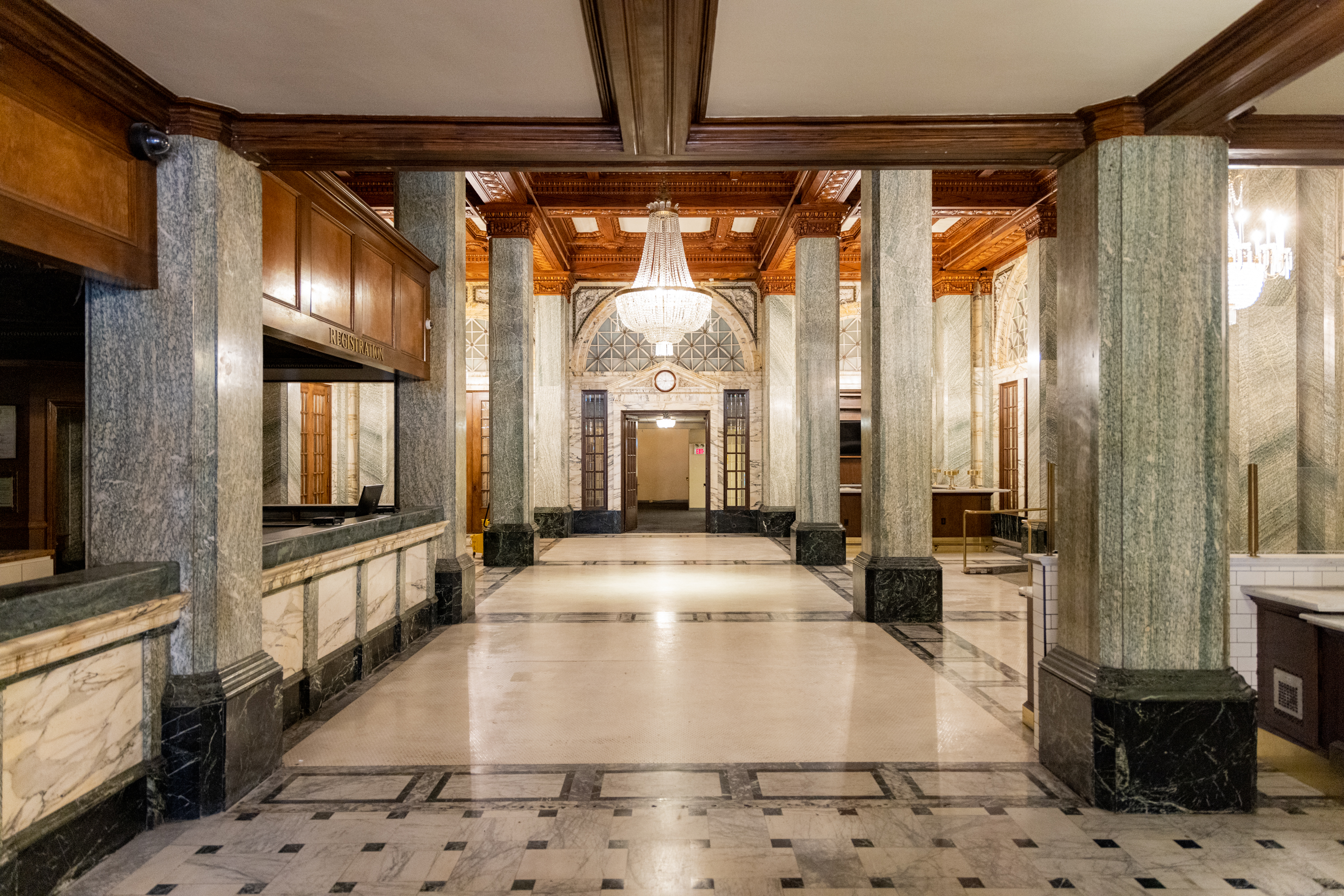 The image shows an elegant lobby with marble floors, tall pillars, wooden accents, and two grand chandeliers. There's a &quot;Registration&quot; sign on a counter.