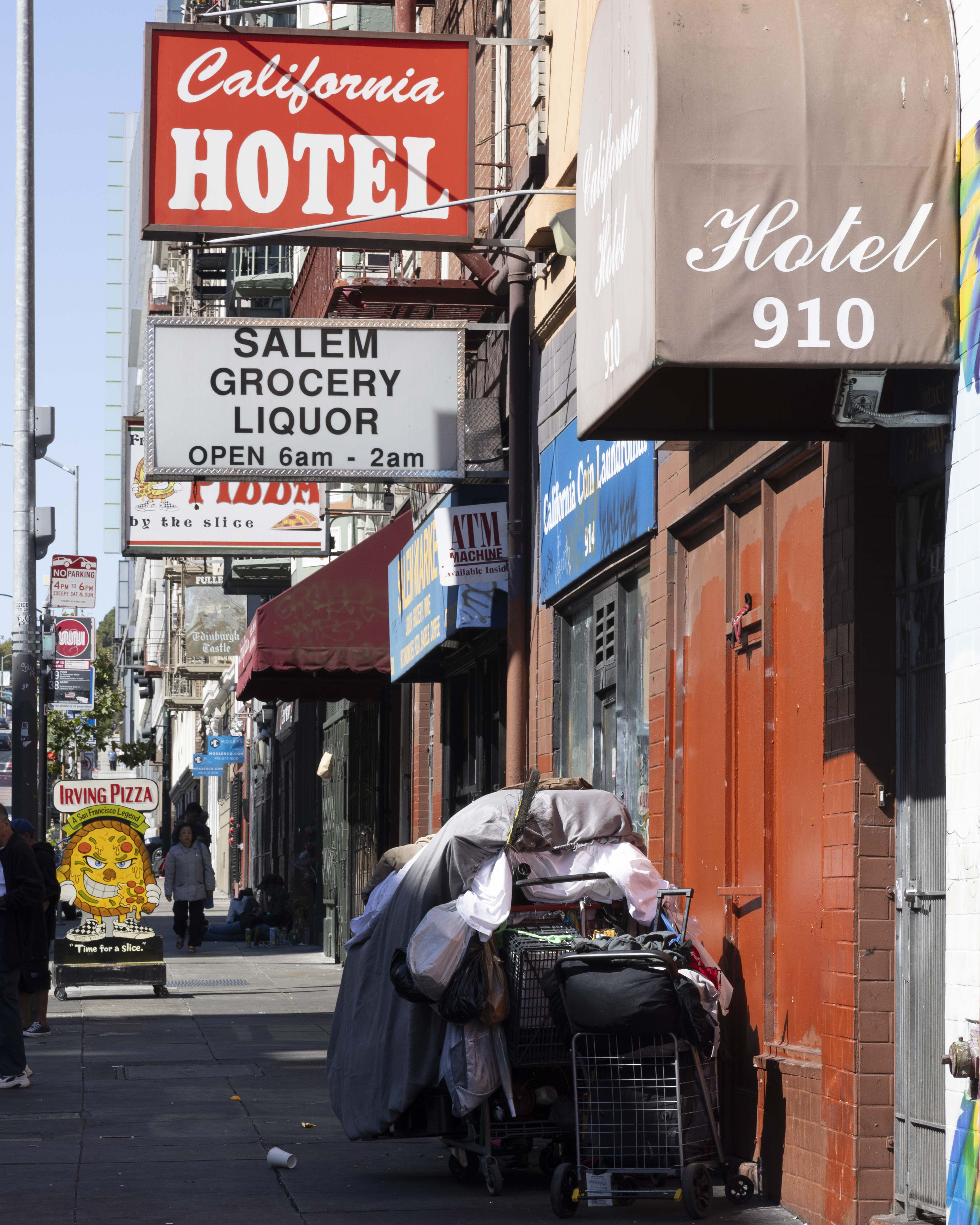 The image shows a busy urban street with a &quot;California Hotel&quot; sign, adjacent stores, and a person pushing a cart laden with belongings near the sidewalk.