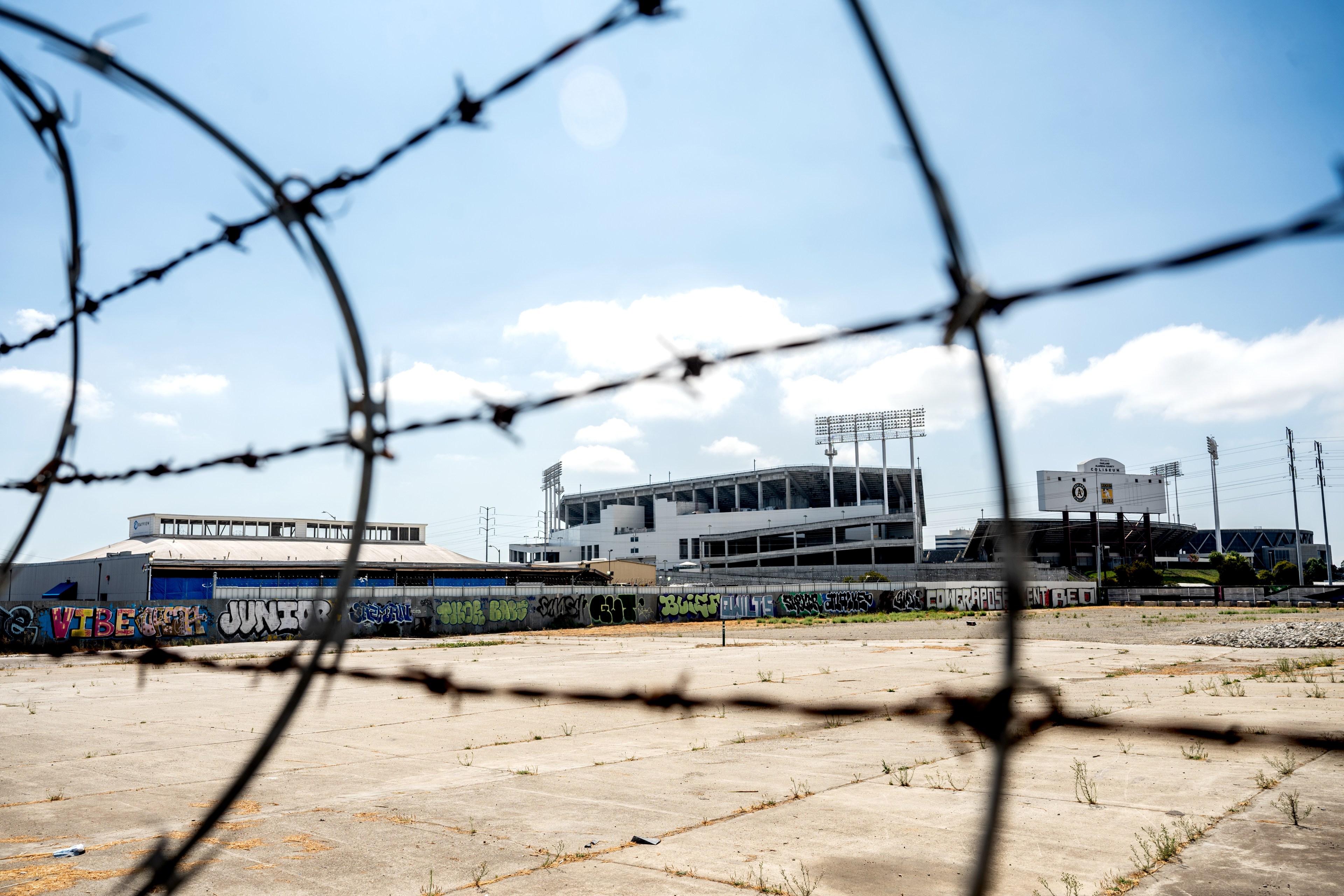 The image shows a stadium behind barbed wire, with graffiti on a concrete wall in the foreground. The sky above is clear with a few clouds.