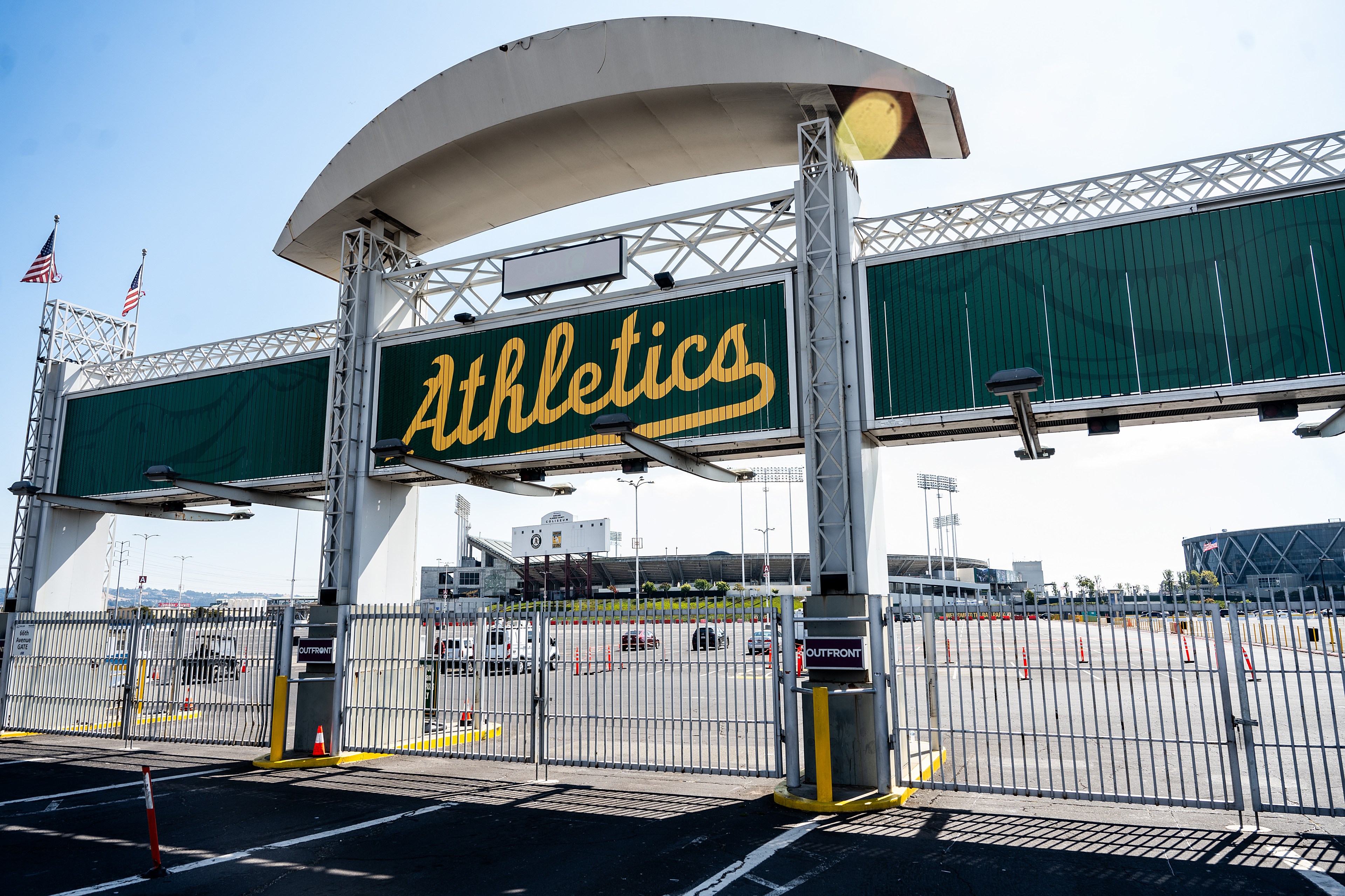 The image shows an entrance gate labeled &quot;Athletics&quot; in yellow script on a green sign, with two American flags above. Beyond the gate is a large, mostly empty parking lot.