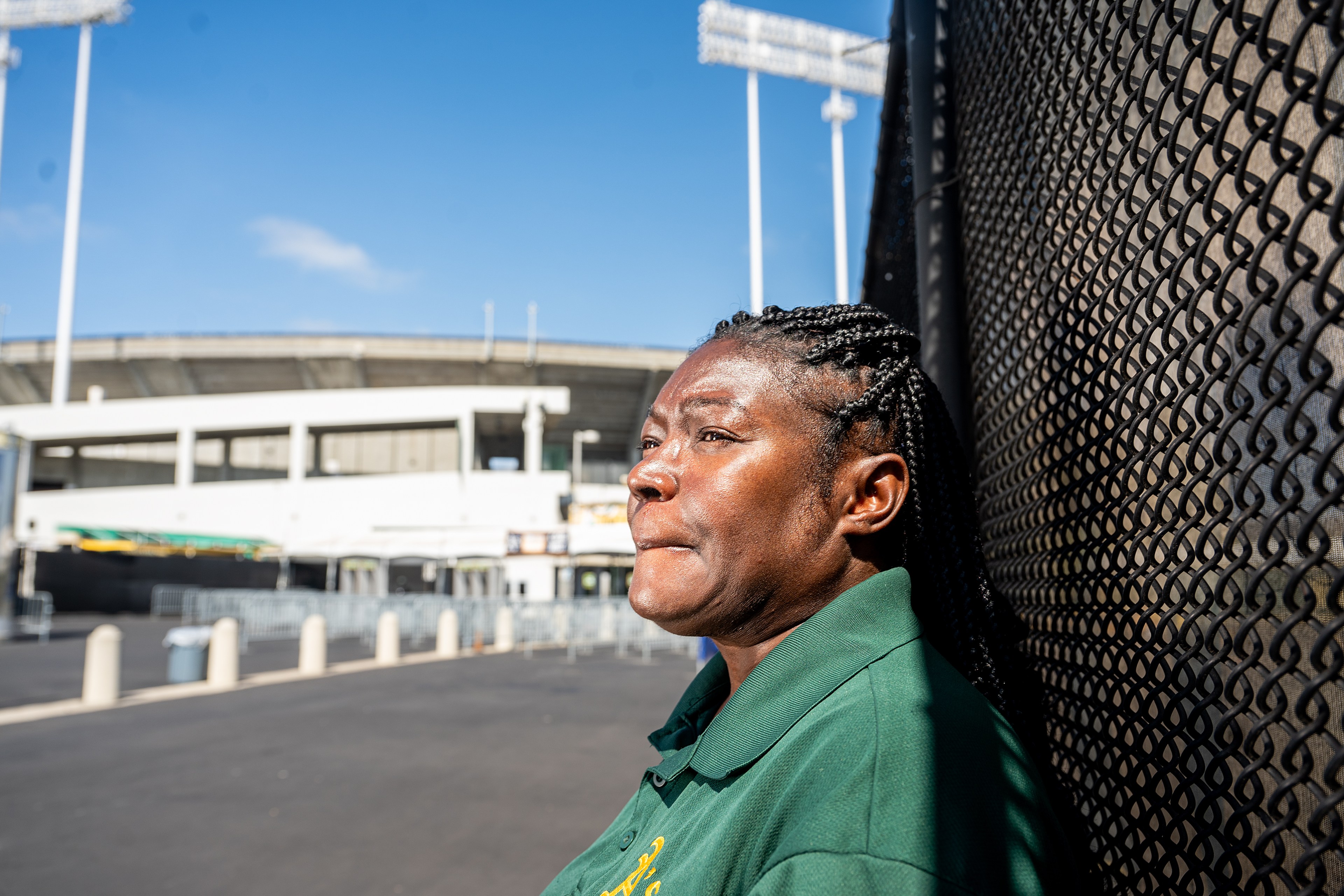 A woman in a green shirt is leaning against a chain-link fence. She's looking into the distance with a thoughtful expression. Behind her is a large stadium.