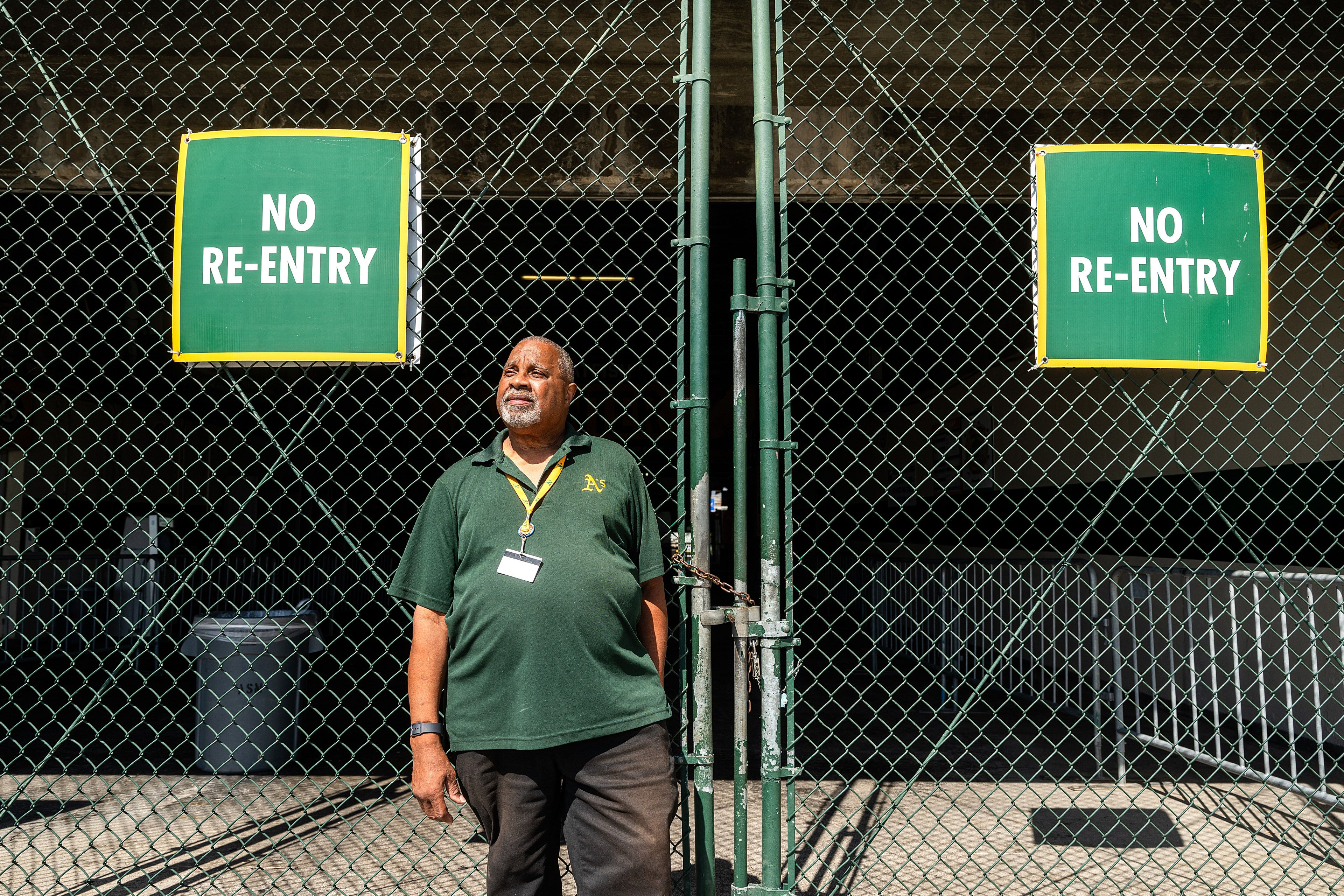 A man in a green shirt stands in front of a chain-link fence with two &quot;No Re-Entry&quot; signs, looking out into the distance.