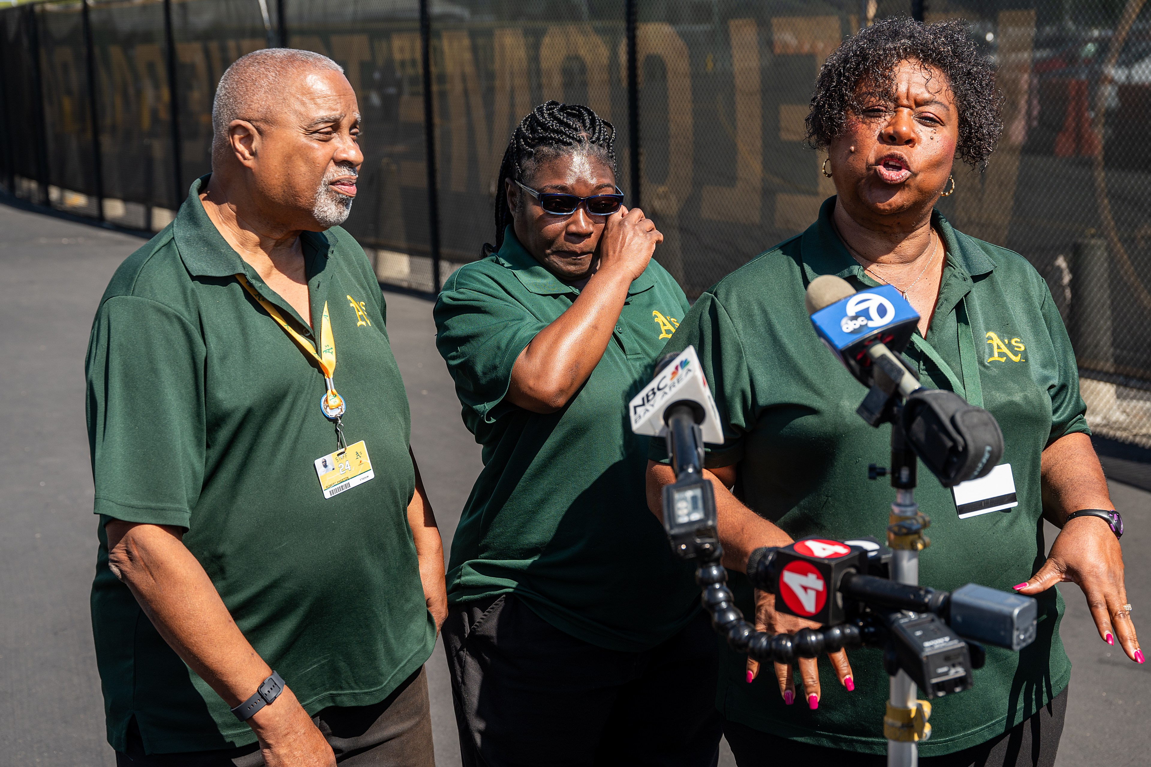 Three individuals in matching green polo shirts stand together outside, two men and a woman, near a chain-link fence. They appear to be addressing multiple news microphones.