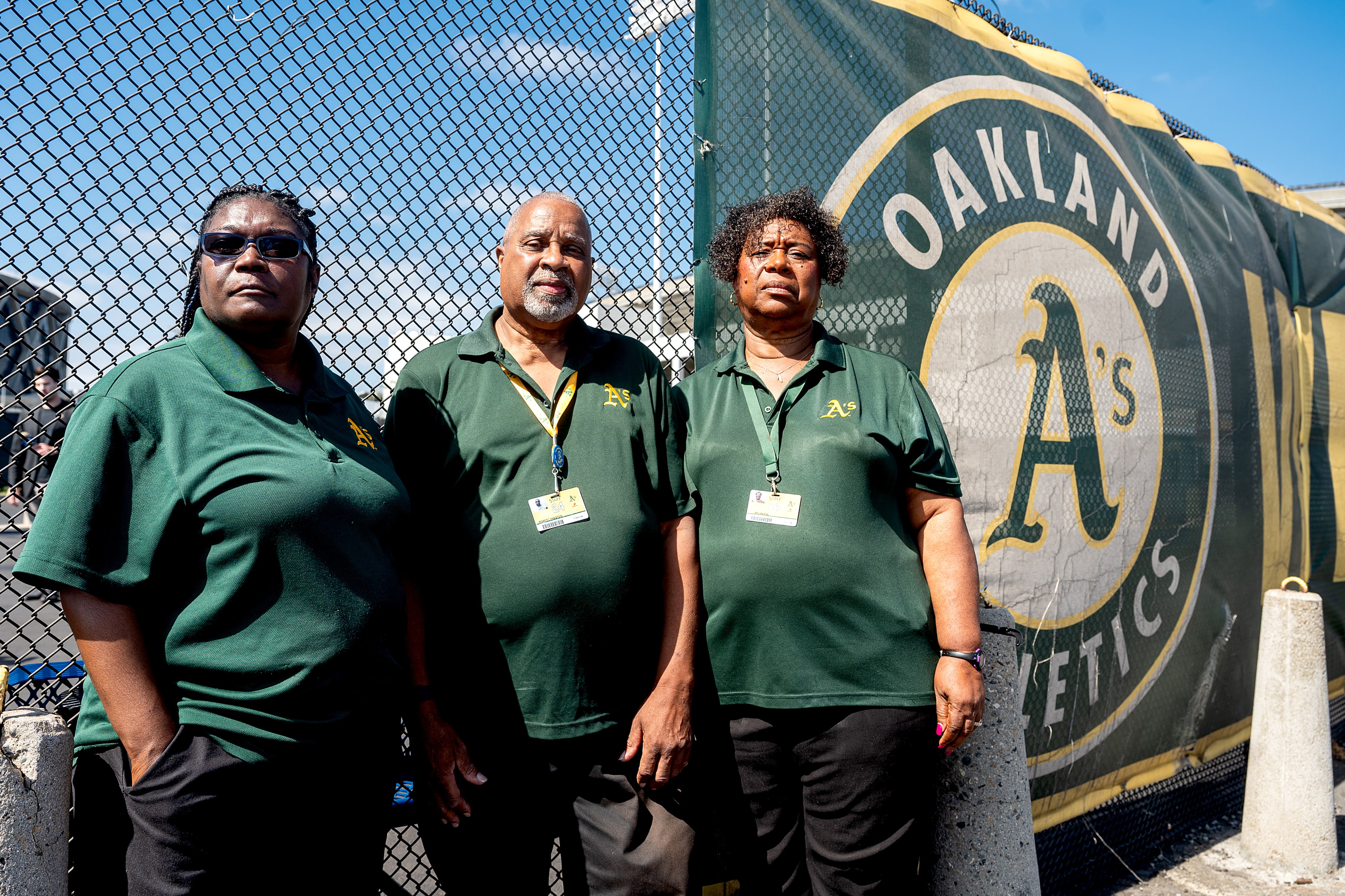 Three people in green shirts and lanyards stand in front of a chain-link fence with an "Oakland Athletics" sign.