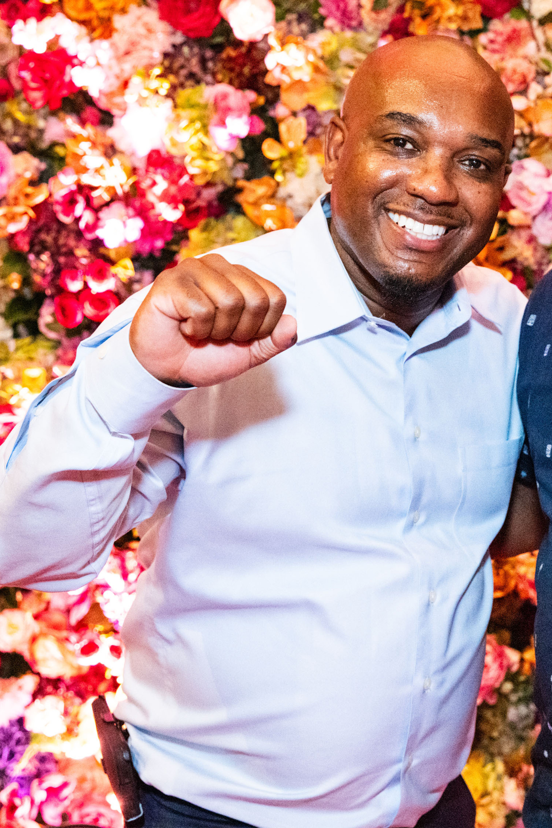 A smiling man in a light blue shirt raises his fist in a friendly gesture against a vibrant, colorful flower wall background.