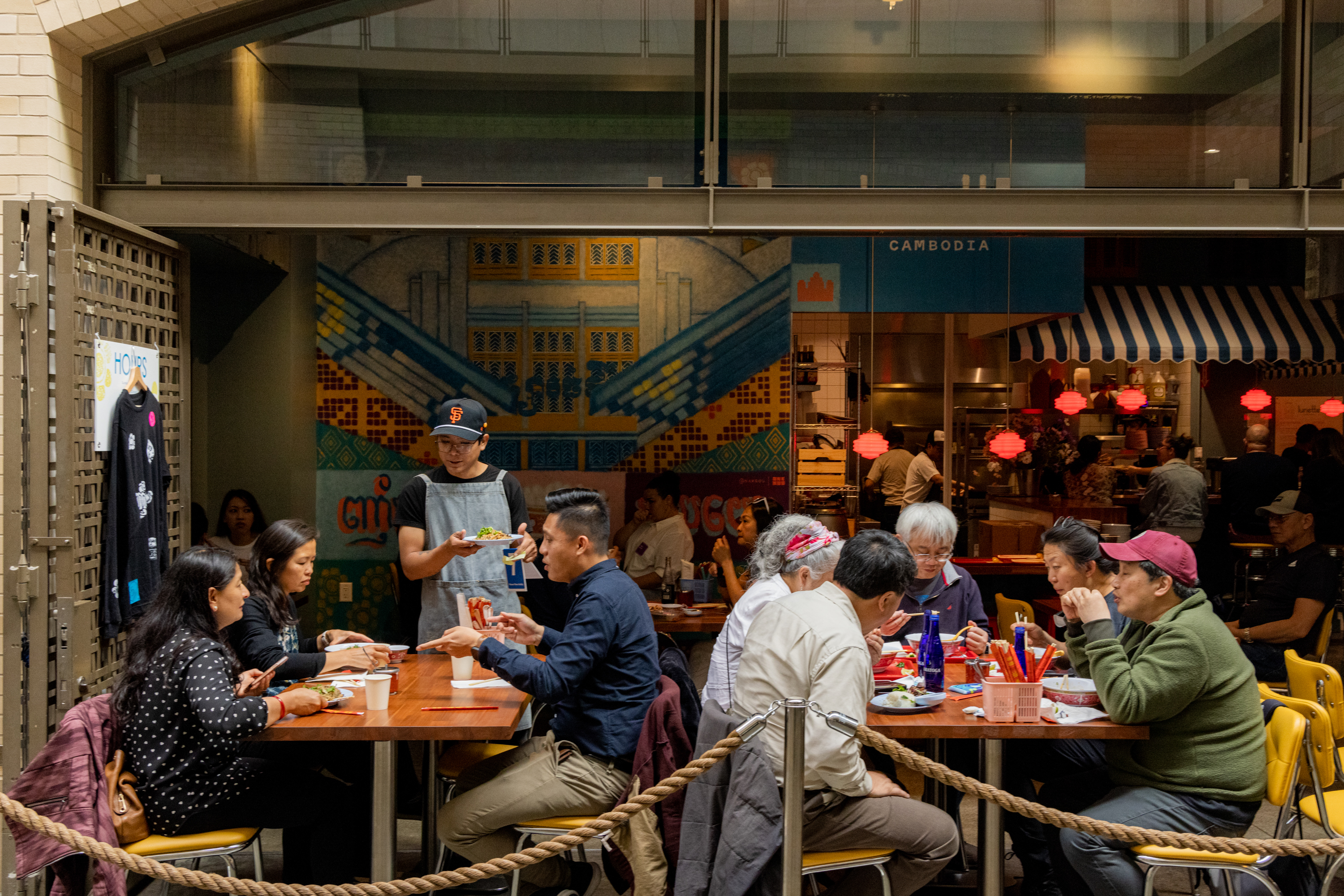 Several people are dining at wooden tables in a vibrant, indoor eatery with colorful decor and a mural in the background. A server is attending to the guests.