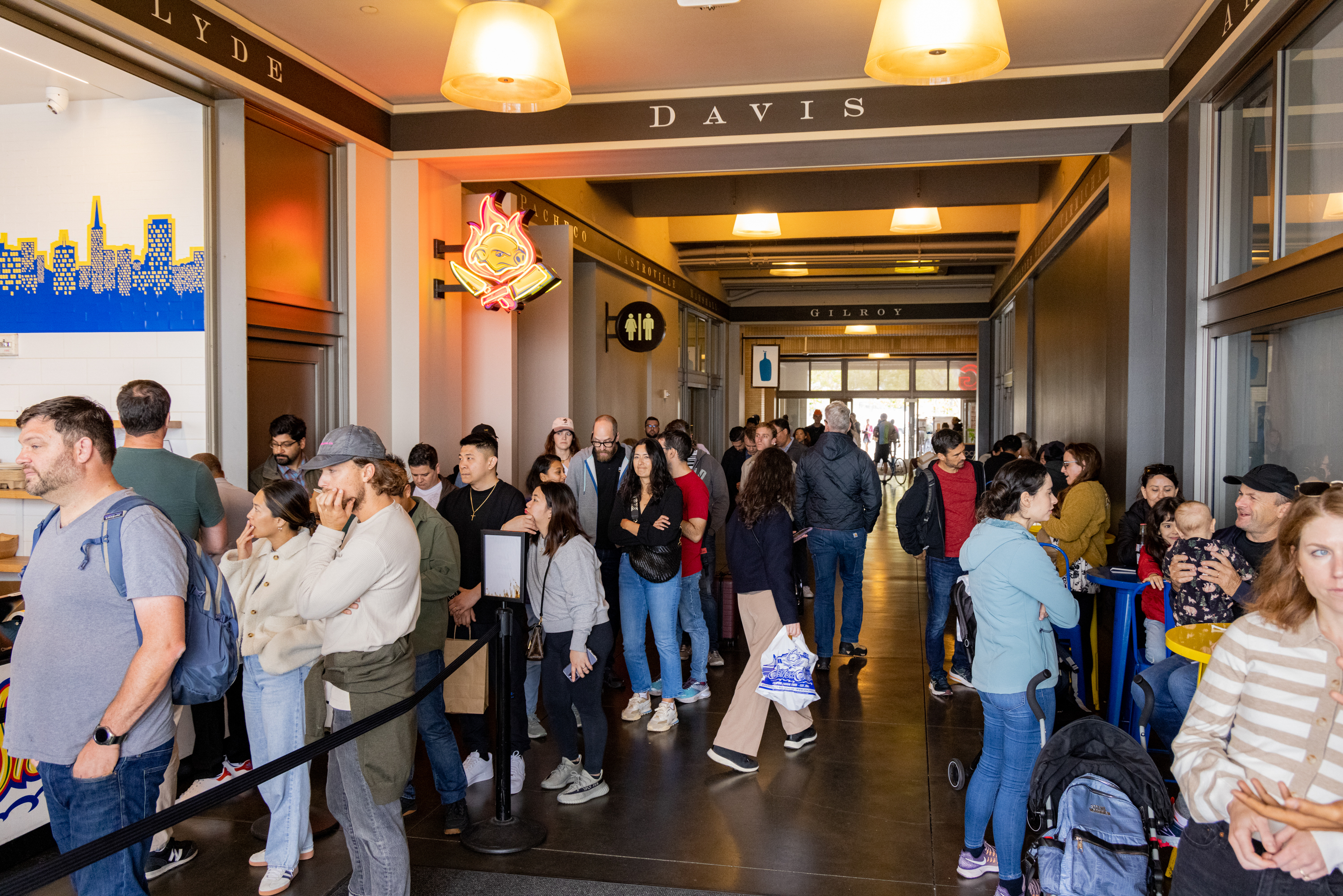 People are queuing in a well-lit hallway with signs above denoting different locations. Some are seated at small tables, while others stand in line chatting or waiting.