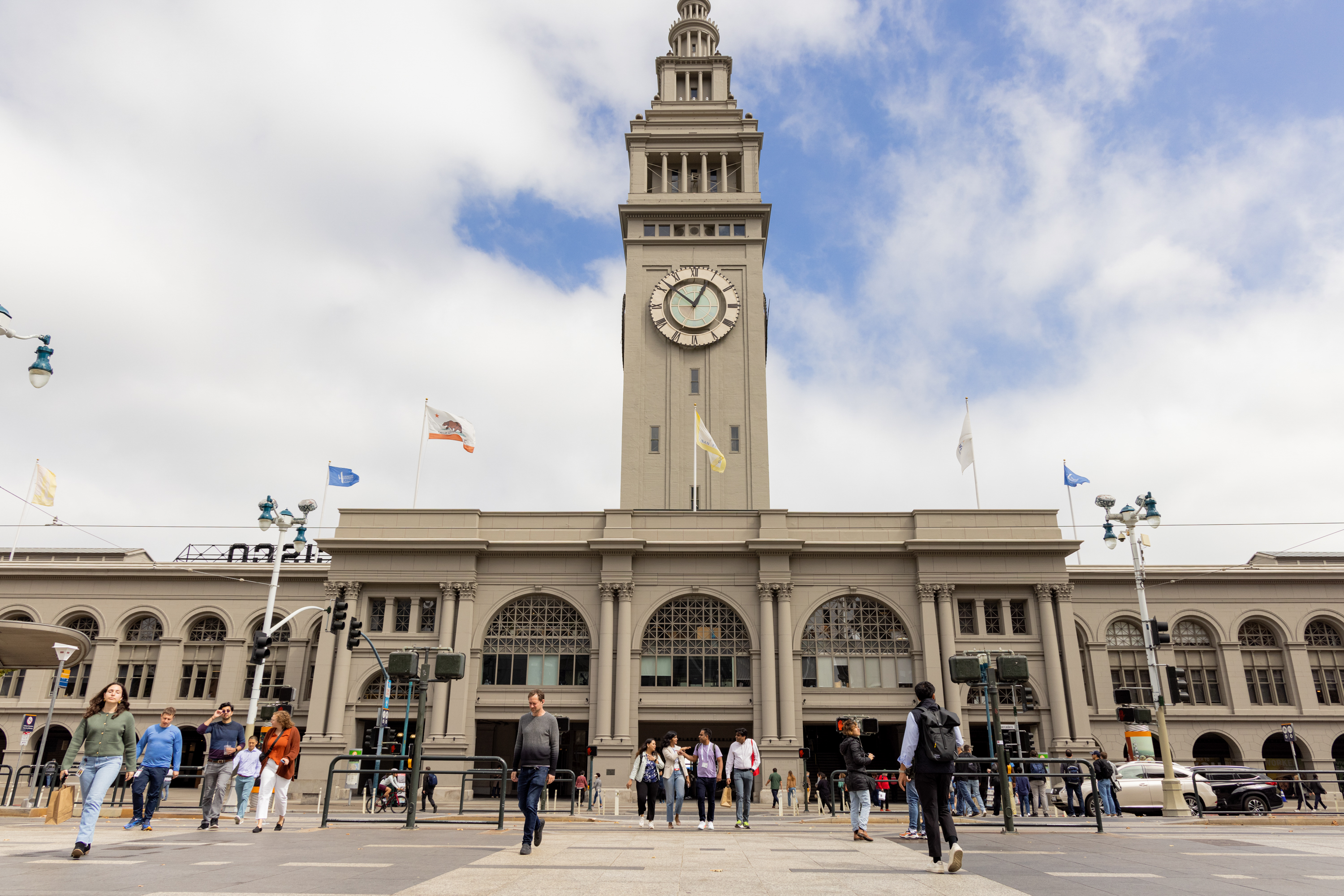 A tall clock tower rises above a gray building with arched windows; people walk in and out of the structure under a partly cloudy sky with flags flying atop the building.