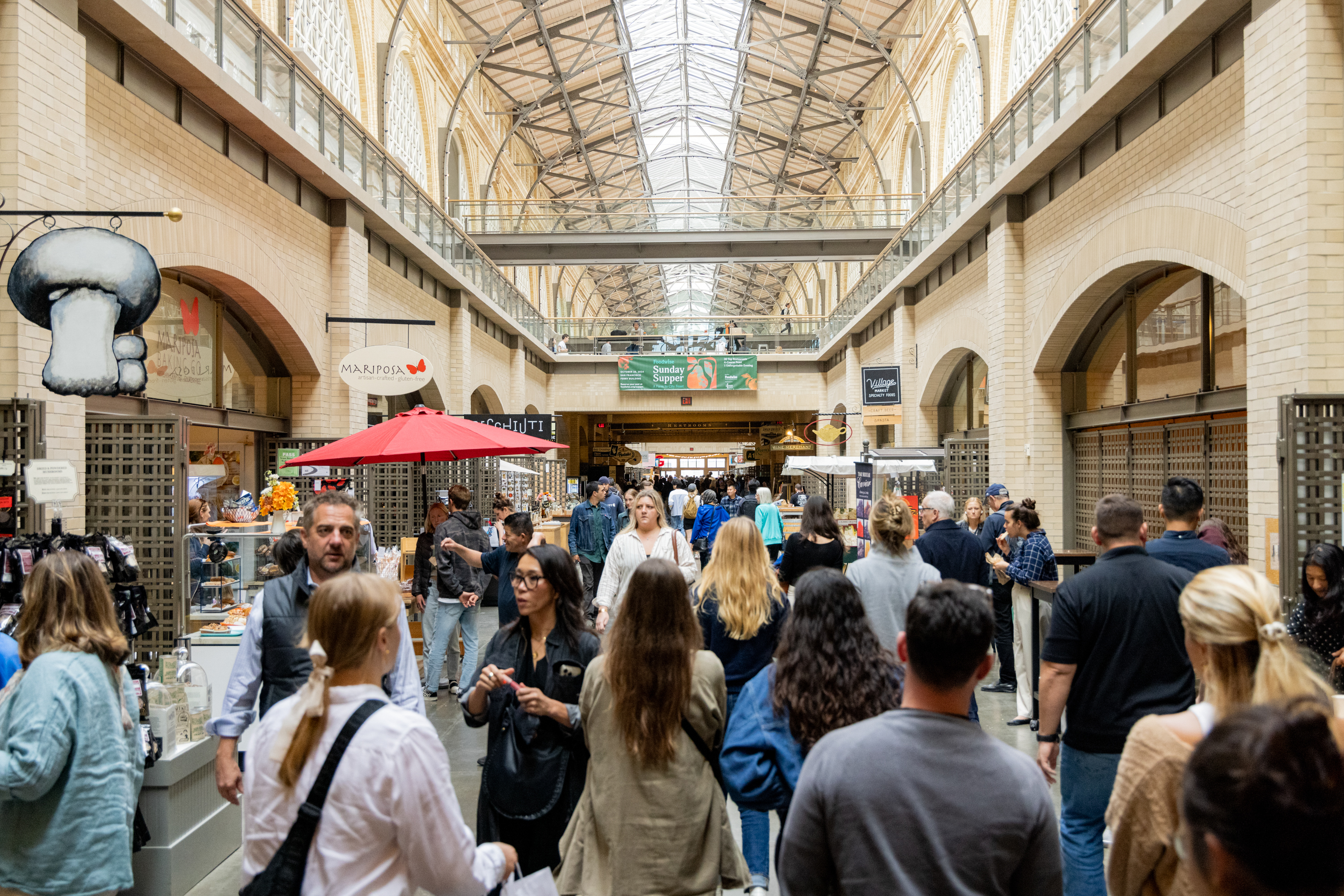 A bustling indoor market with a high arched ceiling, various shop signs, a crowd of people walking, and a red umbrella-covered stall in the center.