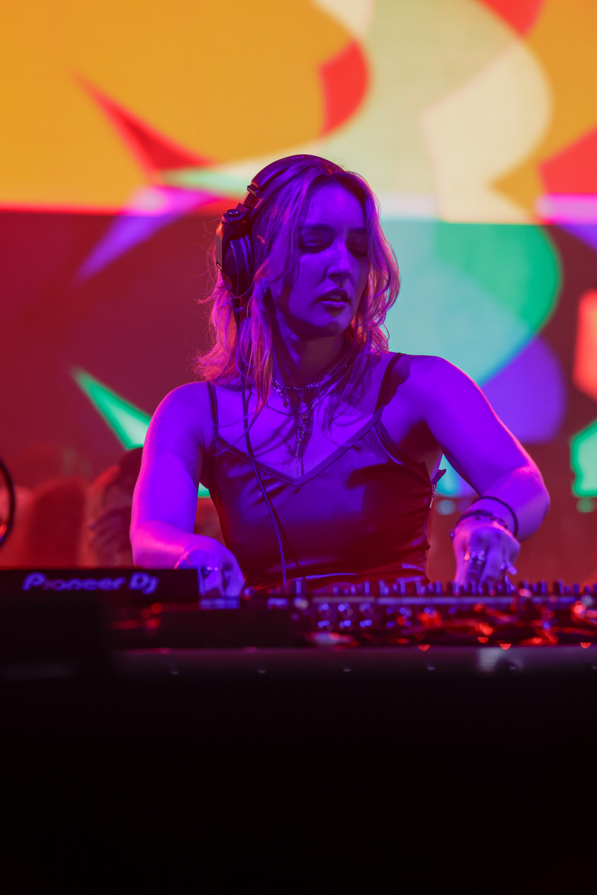 A woman is DJing with headphones on, bathed in purple lighting. She stands behind a mixing console, with a colorful, abstract background behind her.