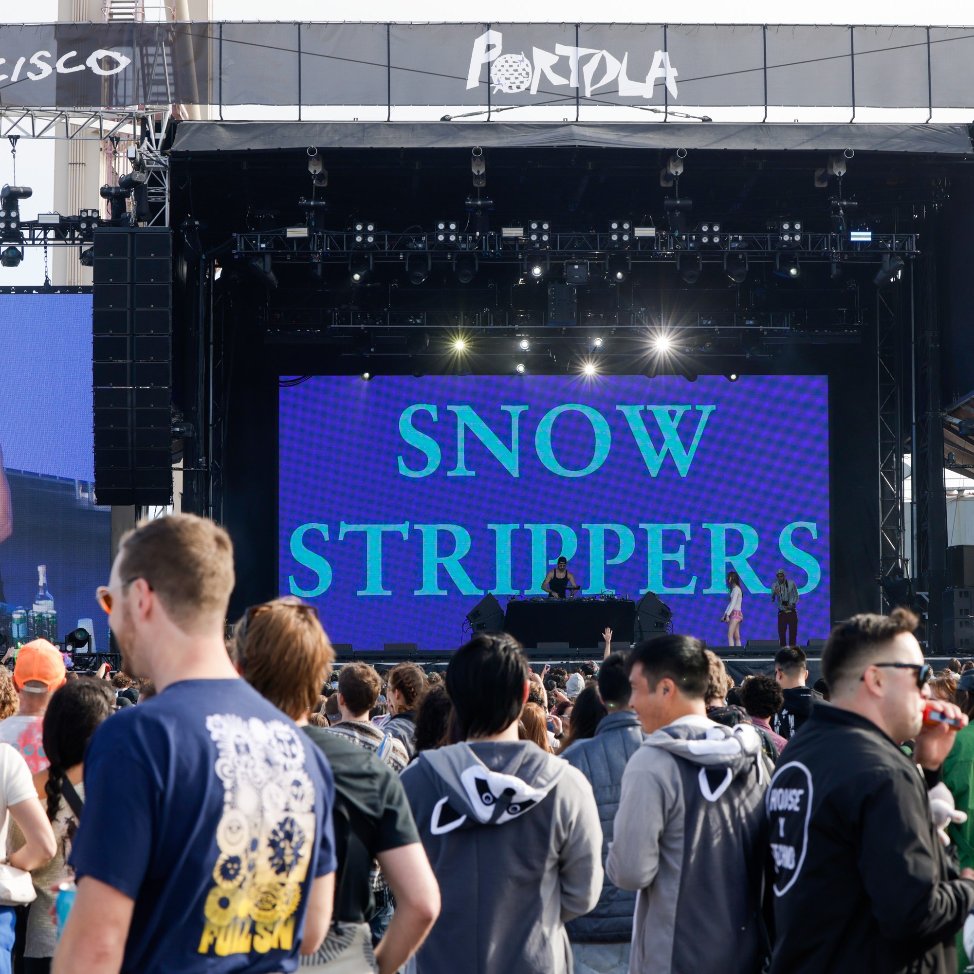 A crowded outdoor concert features a large stage with &quot;Snow Strippers&quot; displayed onscreen. Performers and a DJ are onstage, and the audience is facing them.