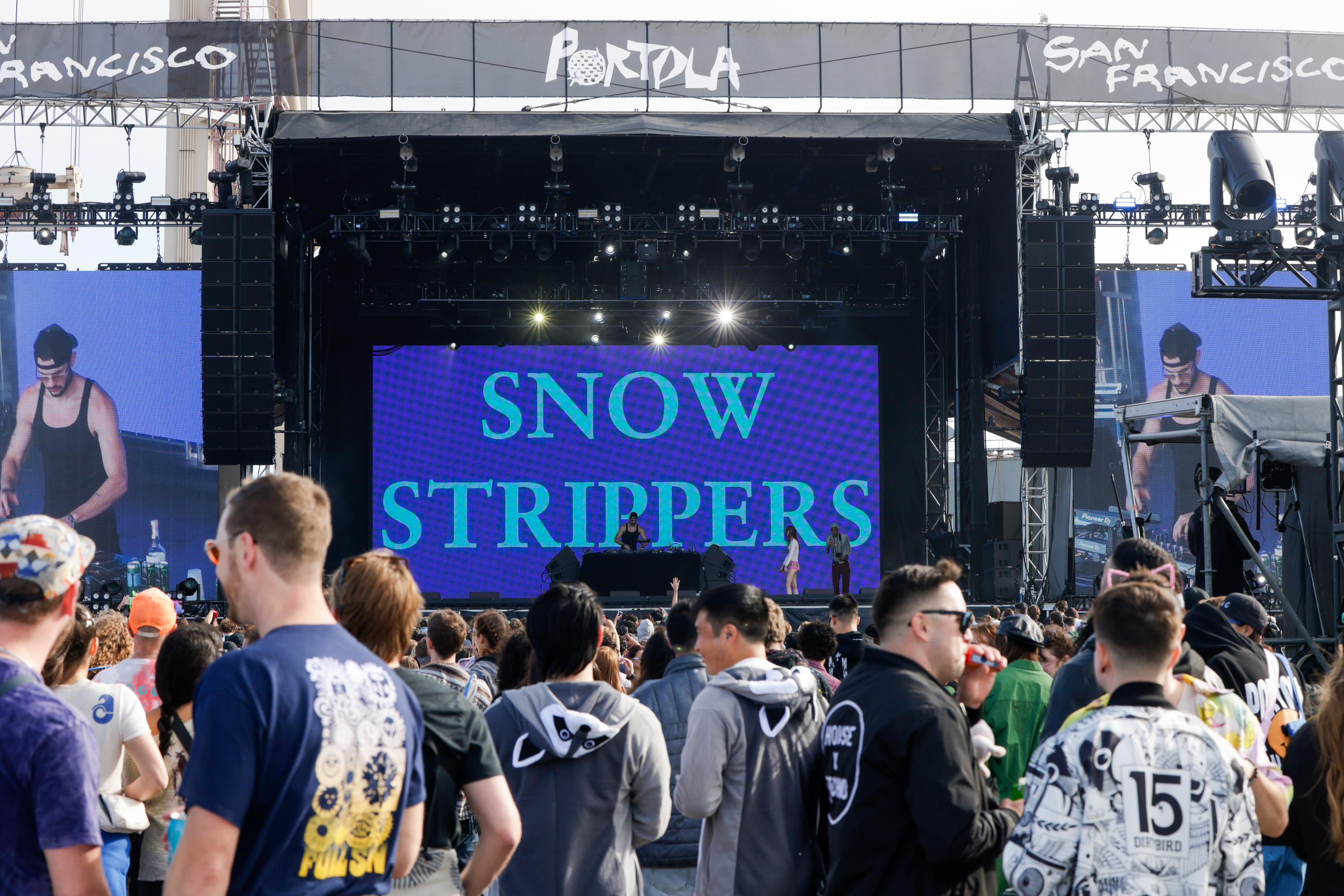 A crowded outdoor concert features a large stage with &quot;Snow Strippers&quot; displayed onscreen. Performers and a DJ are onstage, and the audience is facing them.