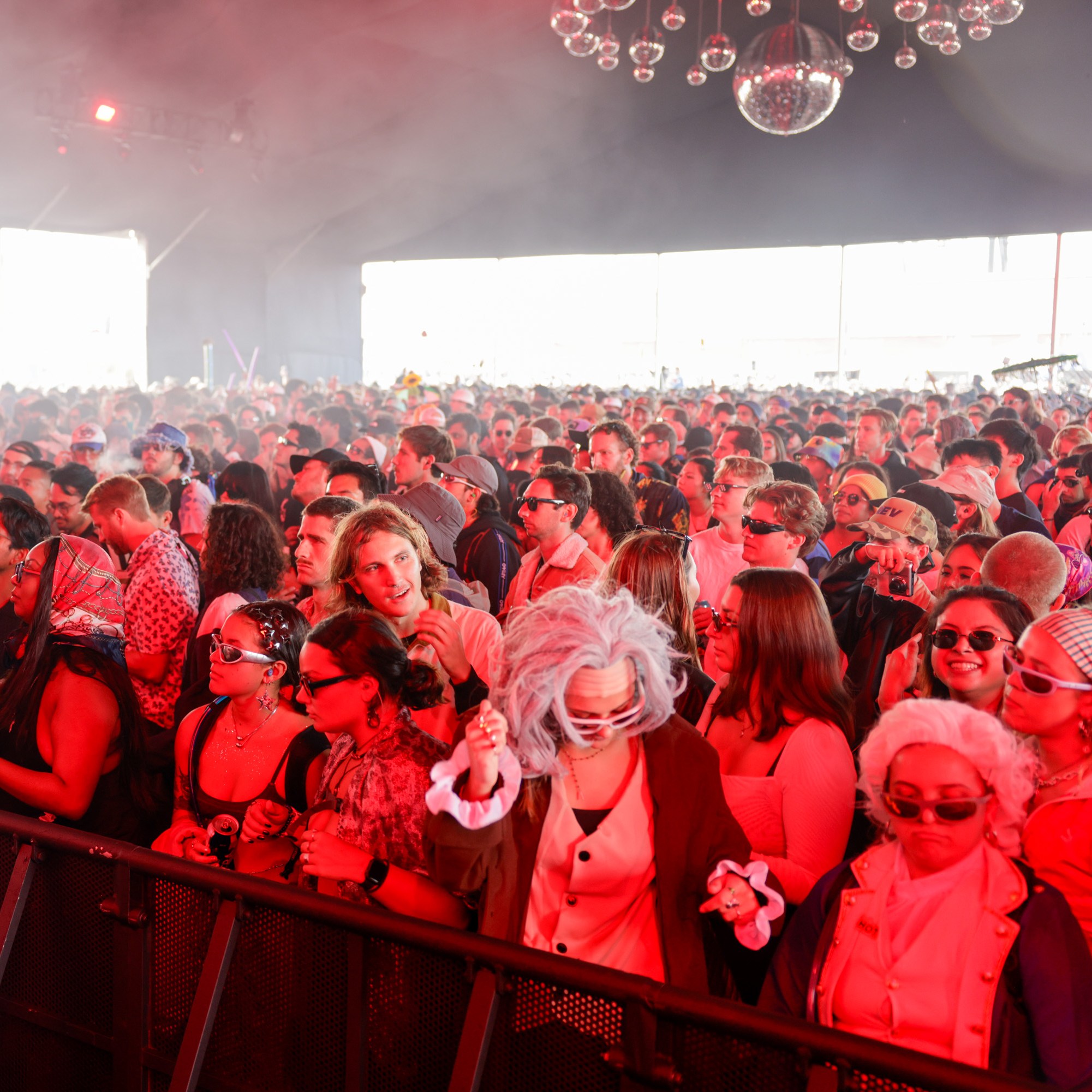 A large, vibrant crowd in costumes and sunglasses is gathered under red lighting at a concert or festival, creating a lively and energetic atmosphere.