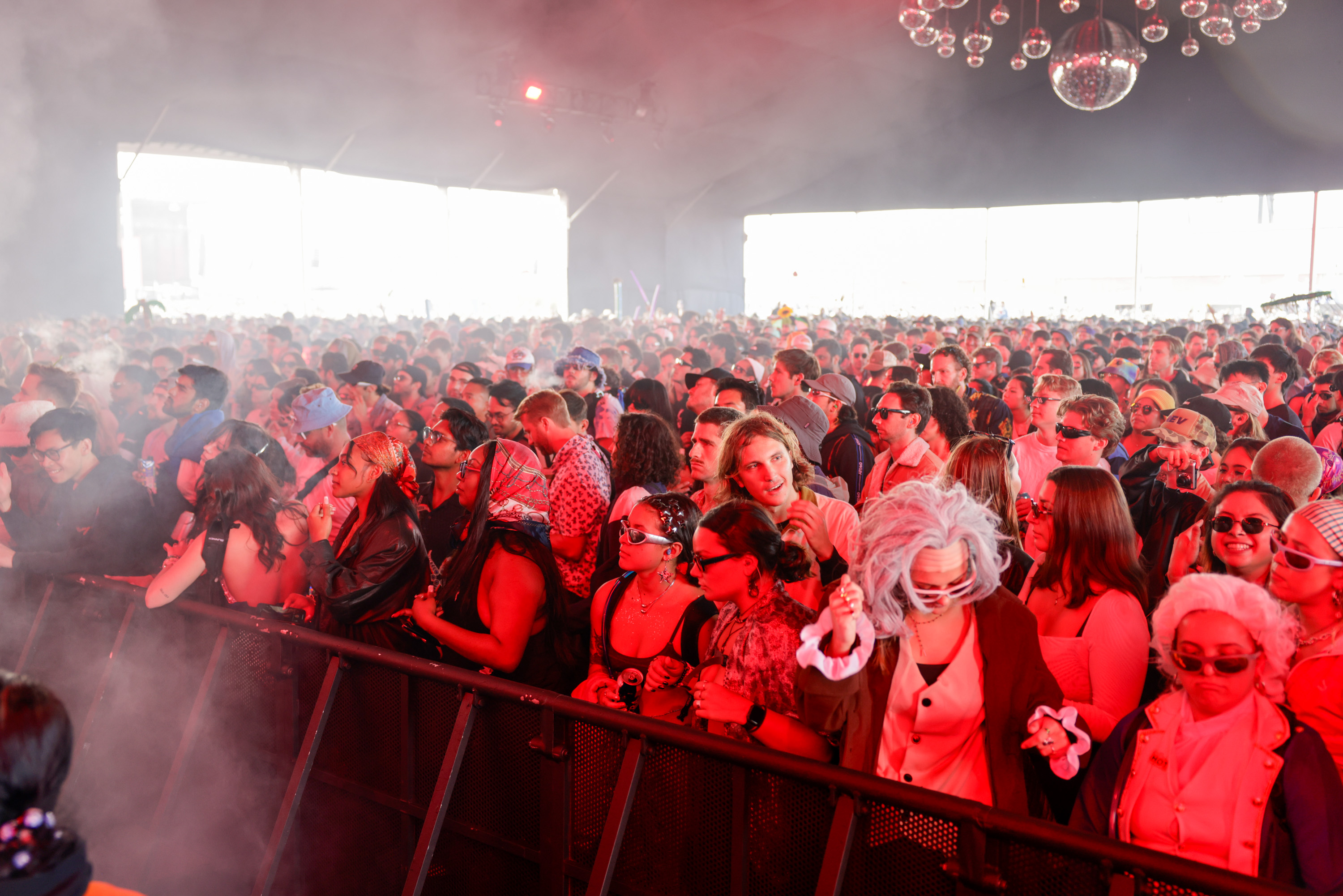 A large, vibrant crowd in costumes and sunglasses is gathered under red lighting at a concert or festival, creating a lively and energetic atmosphere.