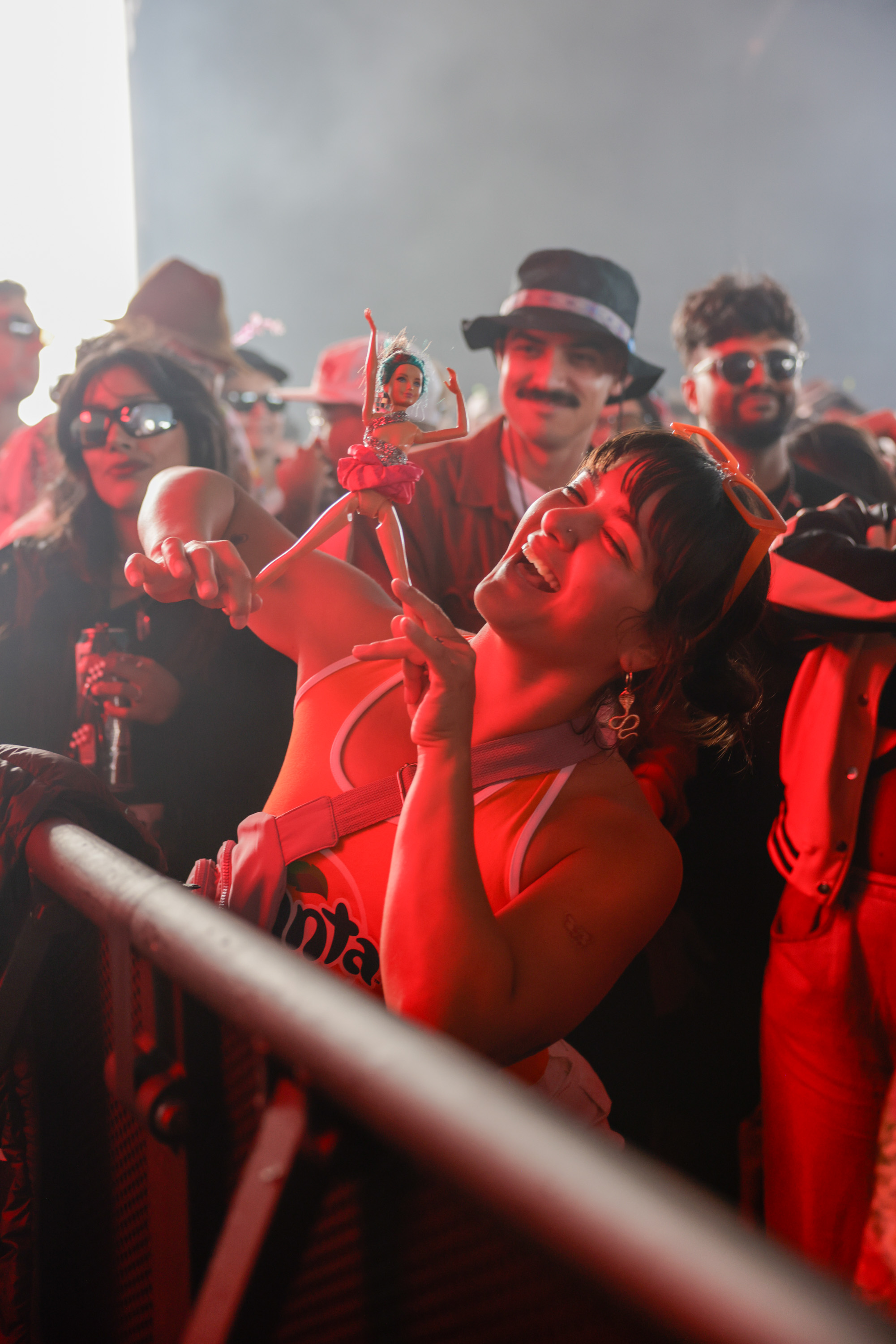 A woman smiling holds a Barbie doll above her head in a crowded, red-lit party, surrounded by other people wearing sunglasses and hats.